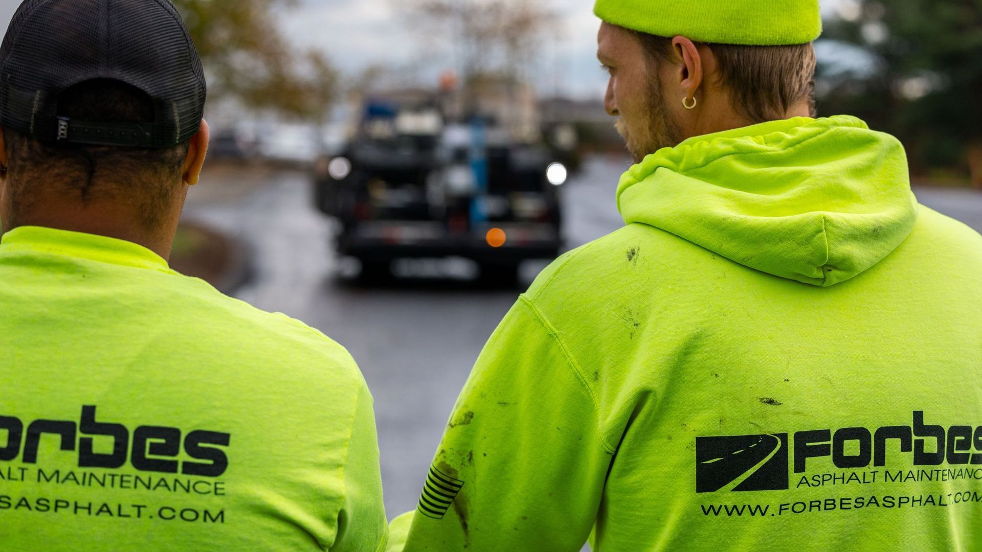 Two men are standing next to each other on a street looking at a car.