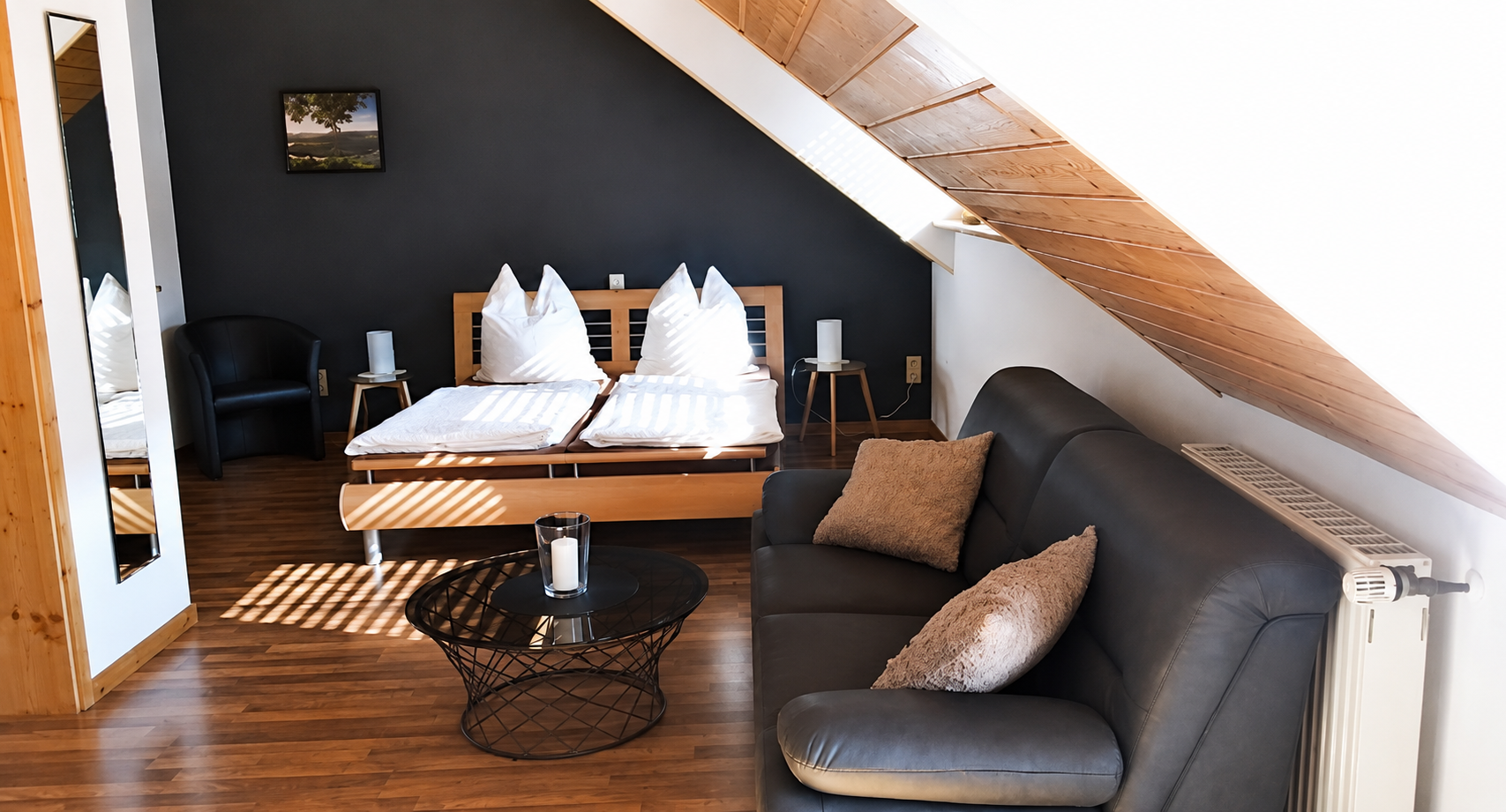 Gemütliches Dachgeschoss-Schlafzimmer mit dunklem Sofa, Bett, Holzbalken und einem kleinen Tisch