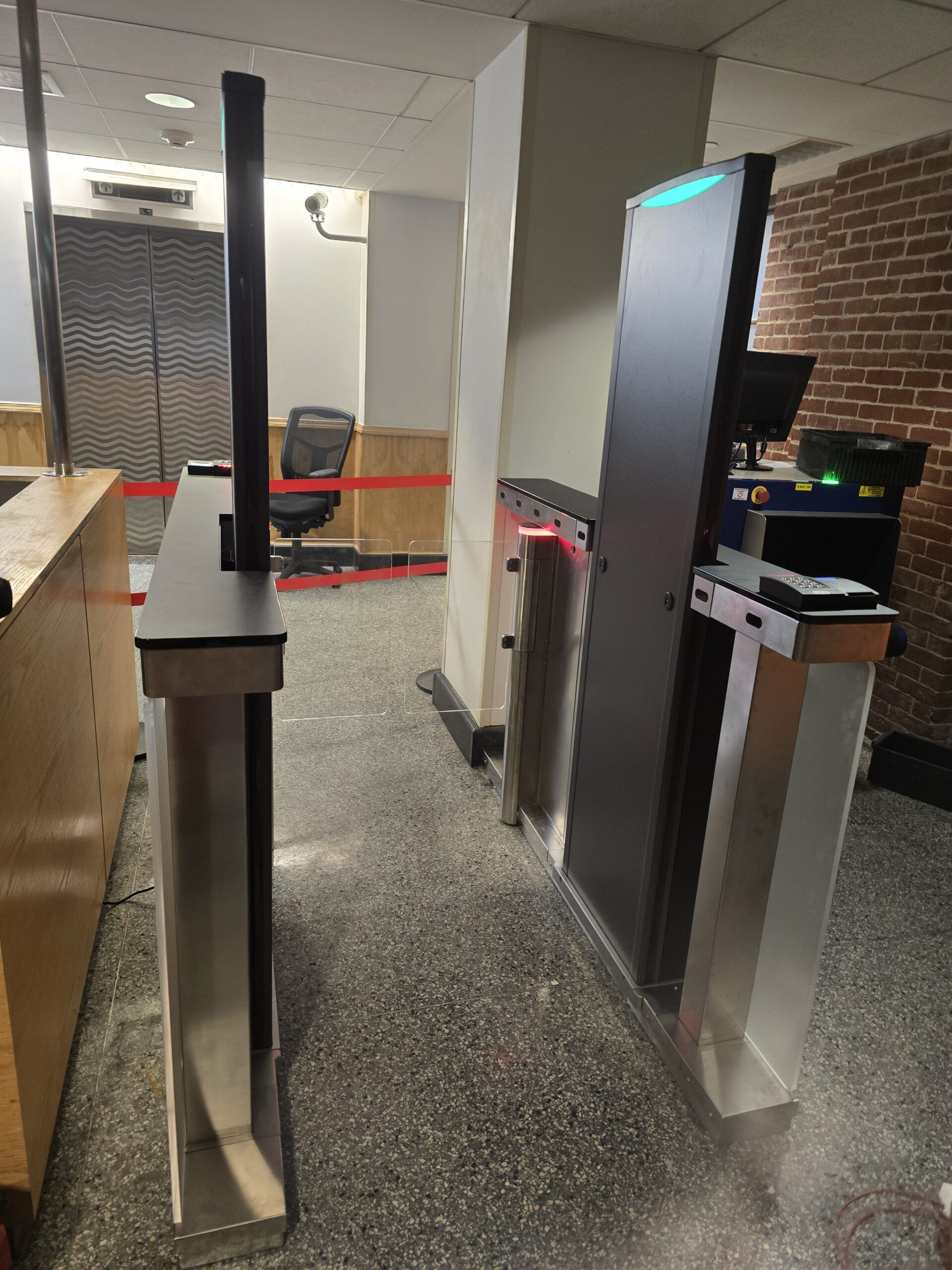 Security turnstile in an office lobby with a desk on the left and a brick wall on the right.