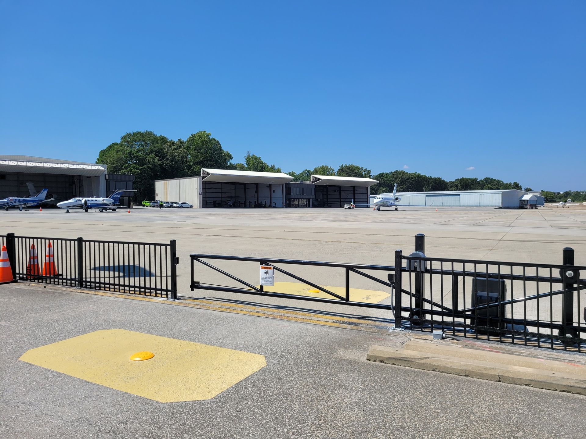 An airfield with airplanes parked near hangars, secured by a gate and fence. Clear, blue sky overhead.