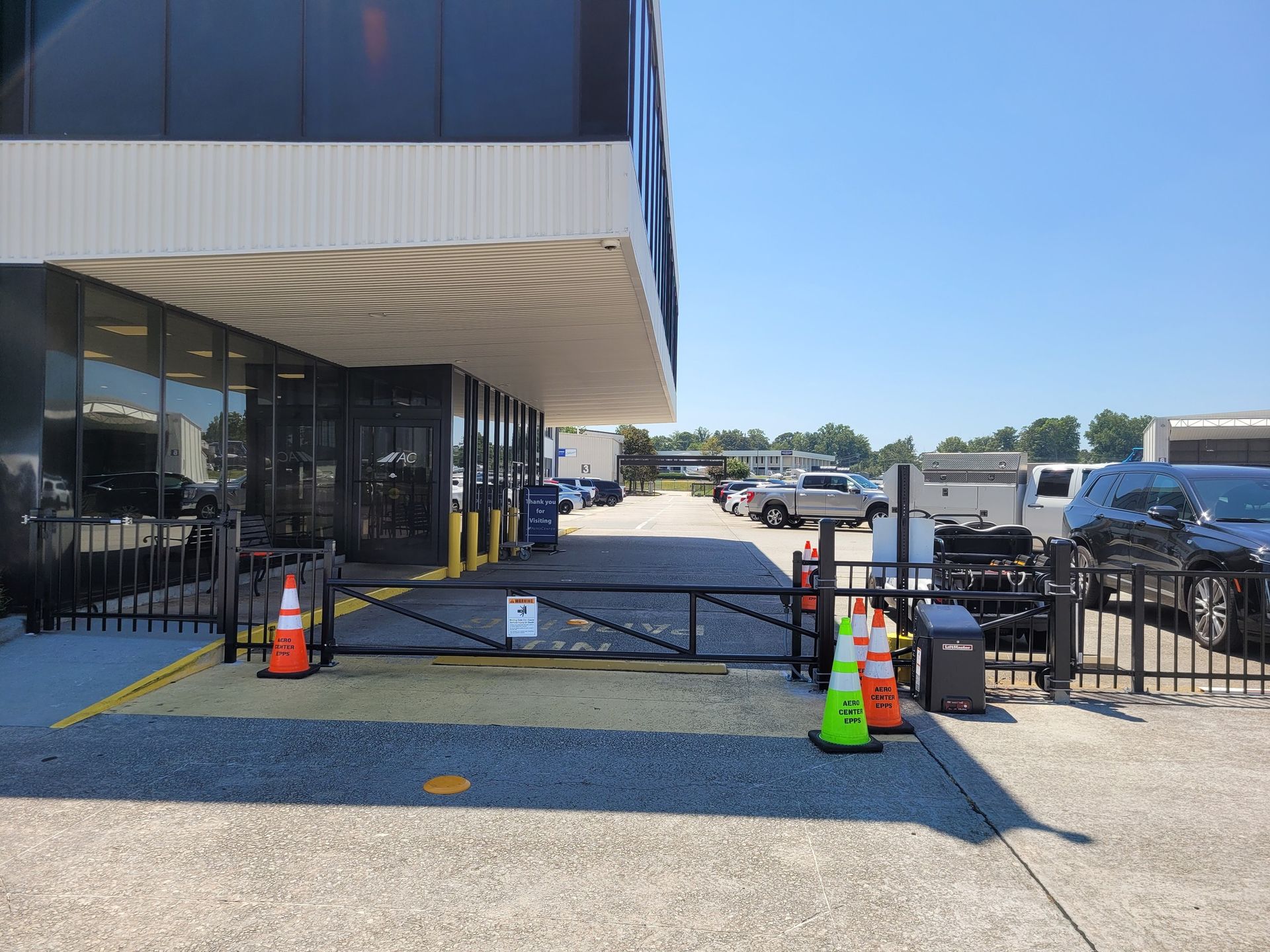 Black gate at building entrance, orange traffic cones, vehicles in parking area, clear sky.