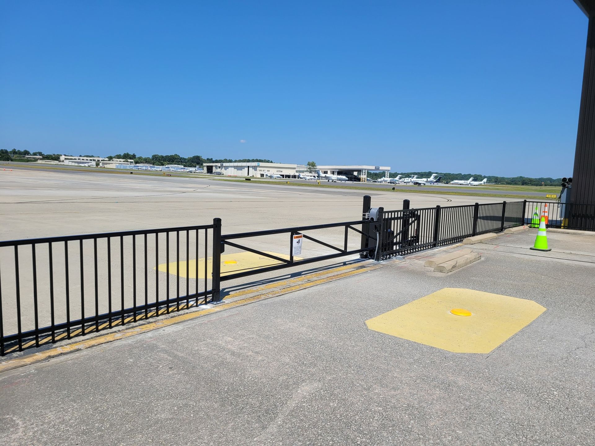 View of an airport tarmac from behind a black metal fence and concrete. A green traffic cone is visible.