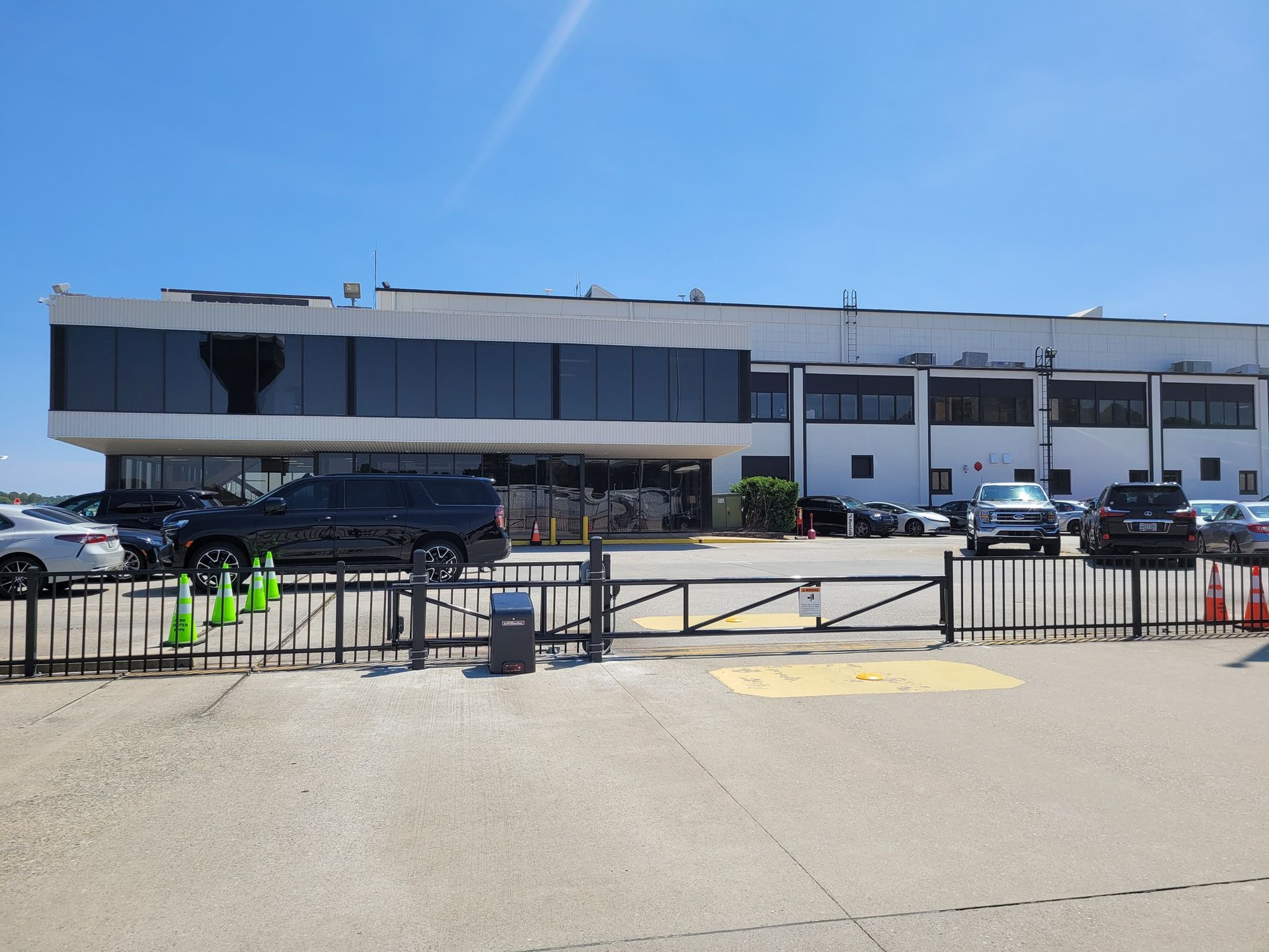 Exterior view of a white building with dark tinted windows. Cars parked in front with a black gate. Bright sunny day.