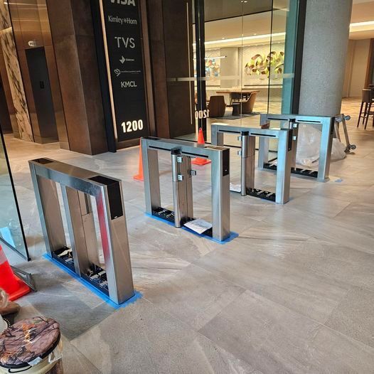 Four silver security turnstiles installed in a building entrance. A sign and seating are visible in the background.
