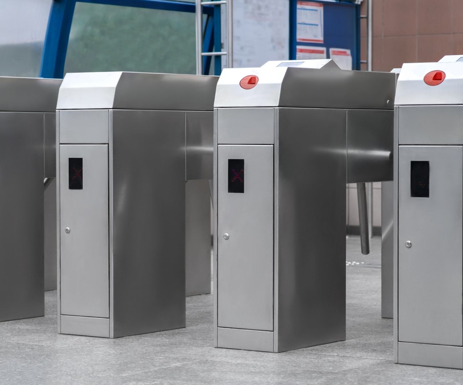 Turnstiles in a transit station. Stainless steel rectangular structures. Red button on top. Dark display panel on front.