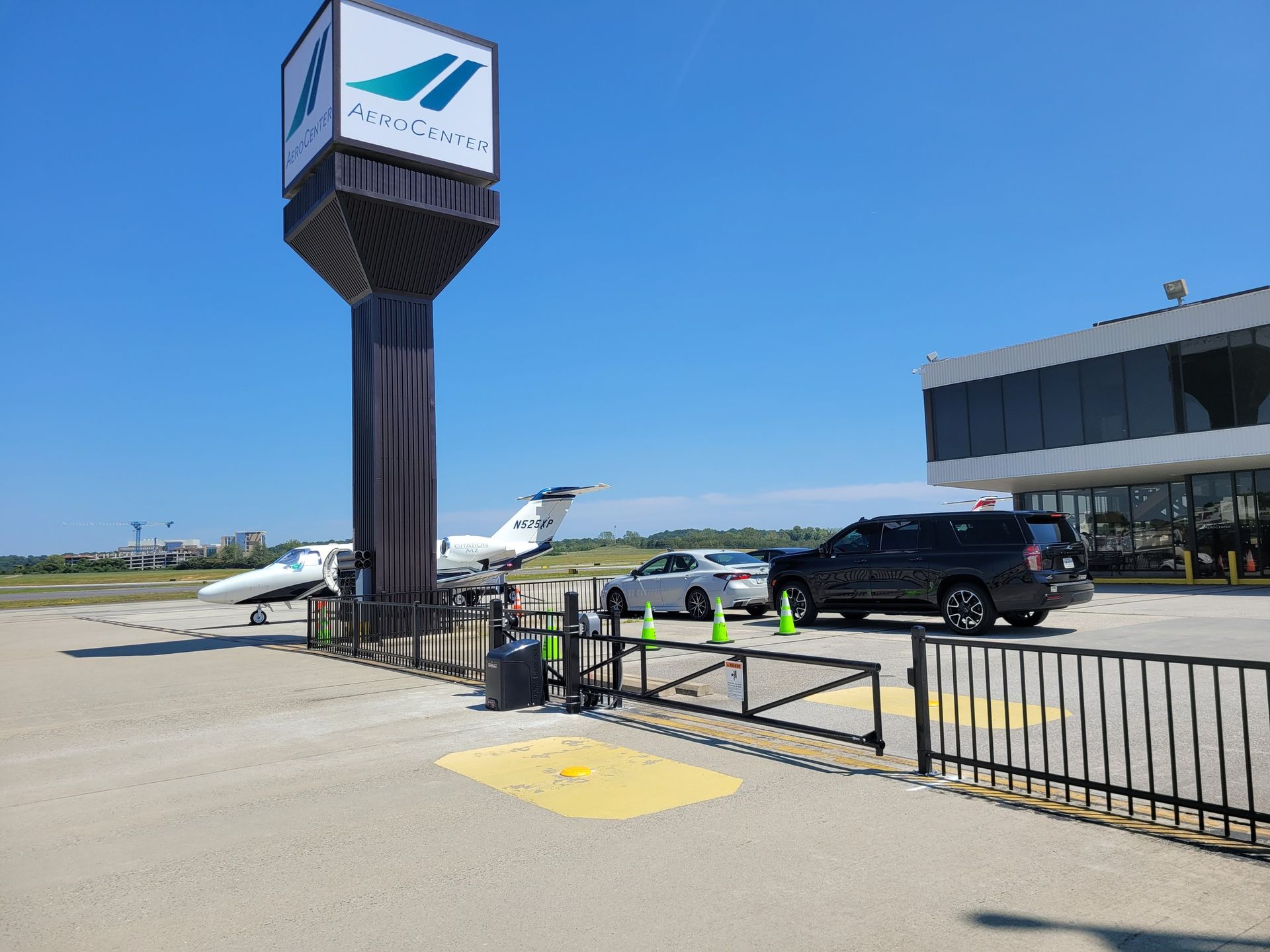 Airport entrance with a sign, planes, cars, and a building on a sunny day.
