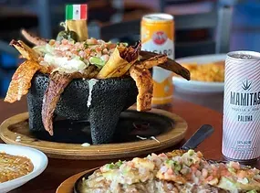 Nachos in a volcanic rock bowl with drinks and food on a table.