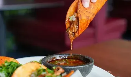 A hand dips a birria taco into a small bowl of consommé; tacos are on a plate.