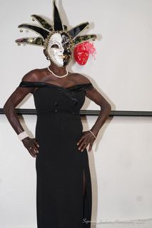 A woman in a black dress and mask is standing with her hands on her hips.