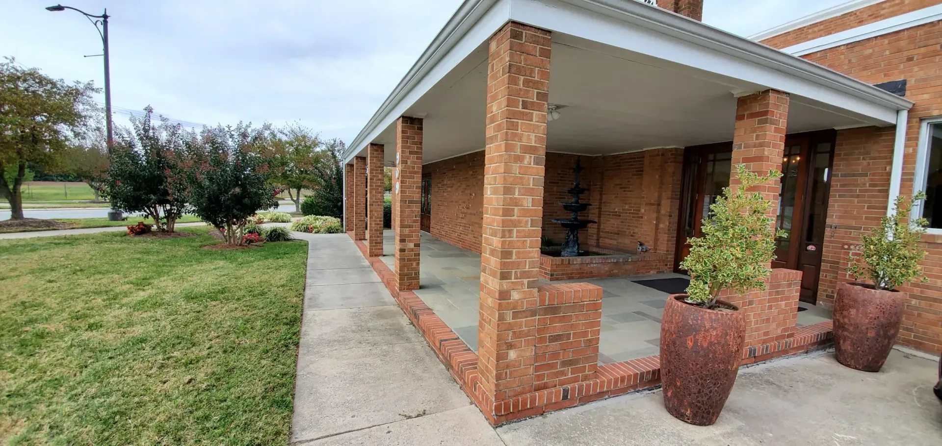 Brick building with porch, brick pillars, plants in large pots. Paved walkway and grassy lawn. Cloudy sky.