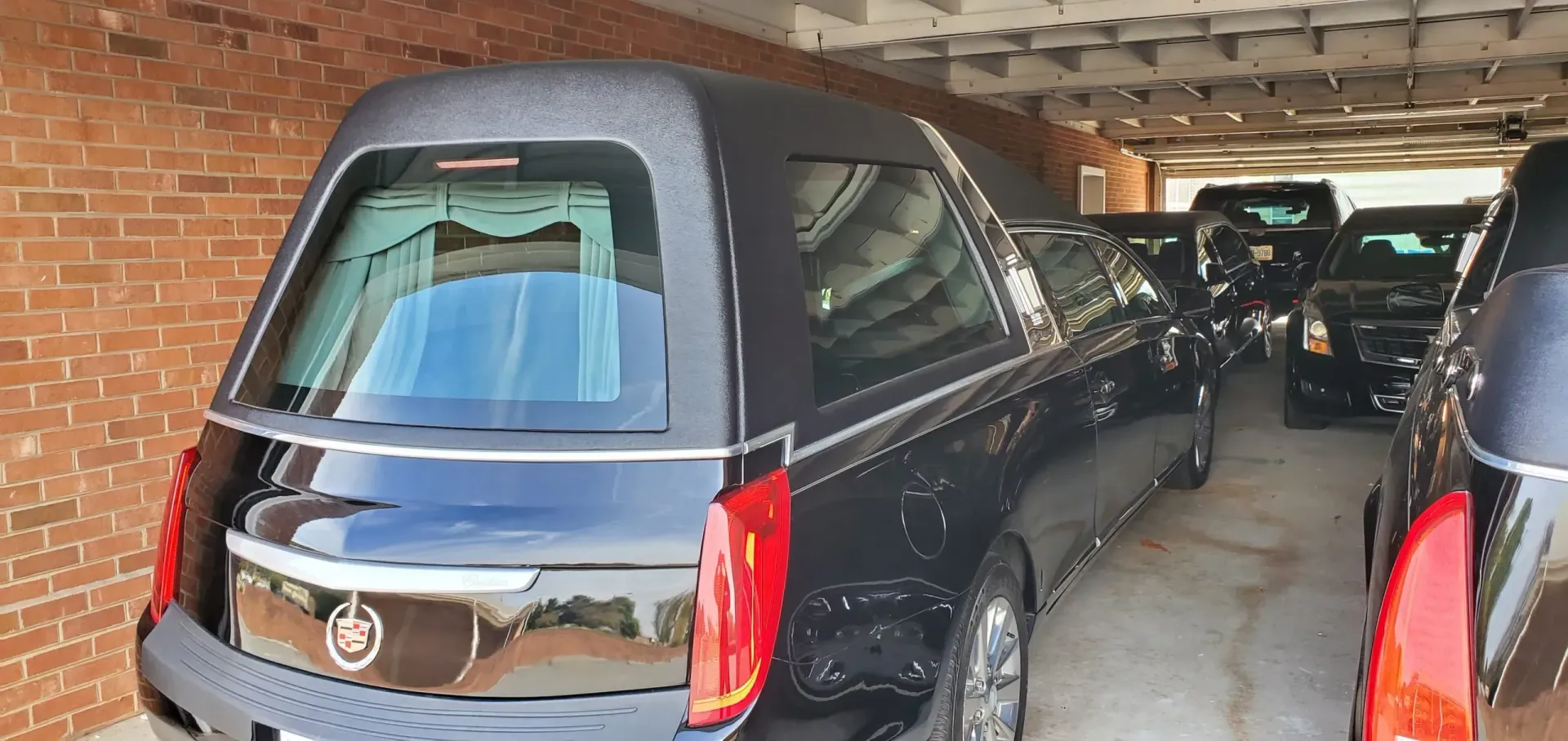 Black hearse parked in a garage with other black hearses. Red brick wall.