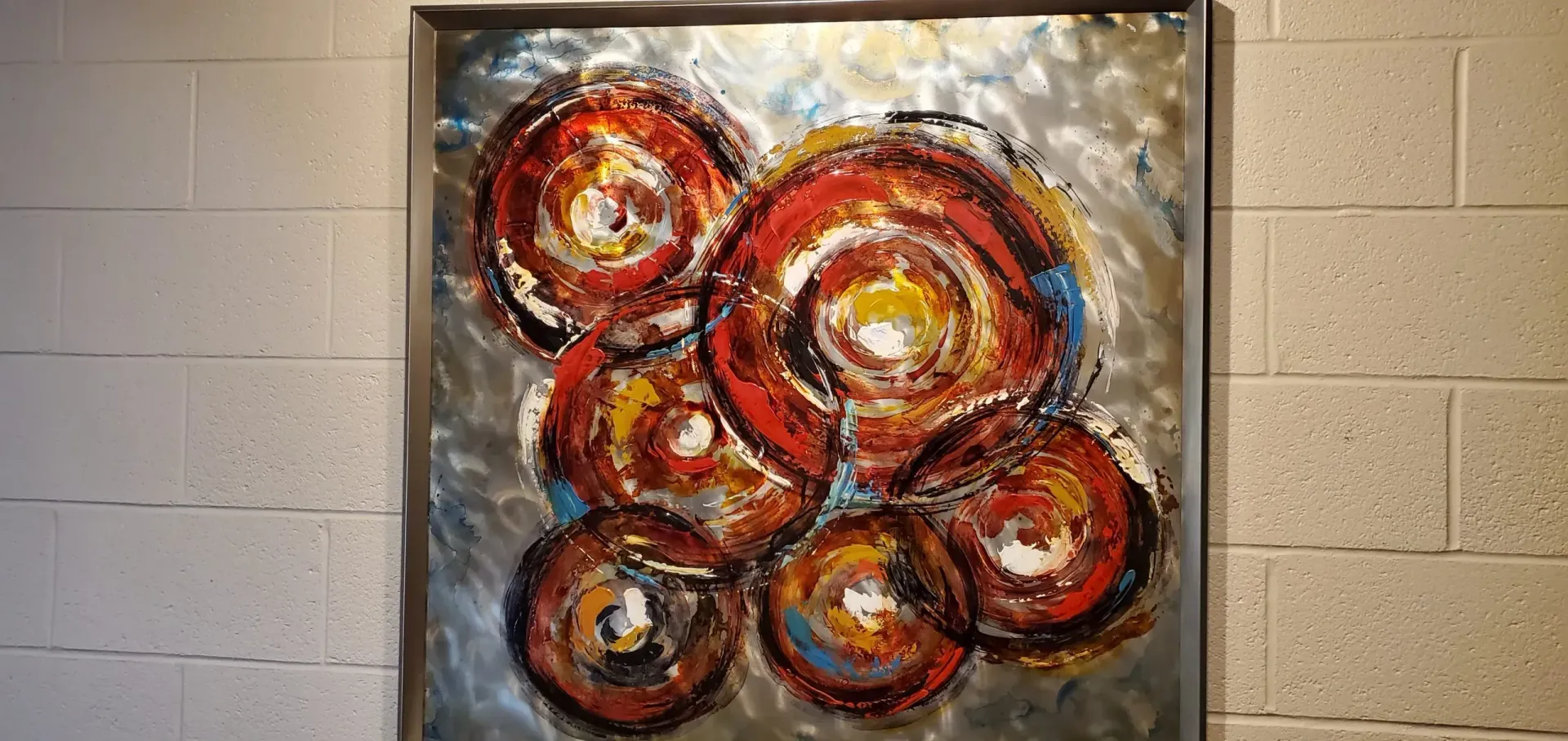 Abstract painting with layered red, orange, and yellow circles on a silver background, in a silver frame, on a brick wall.