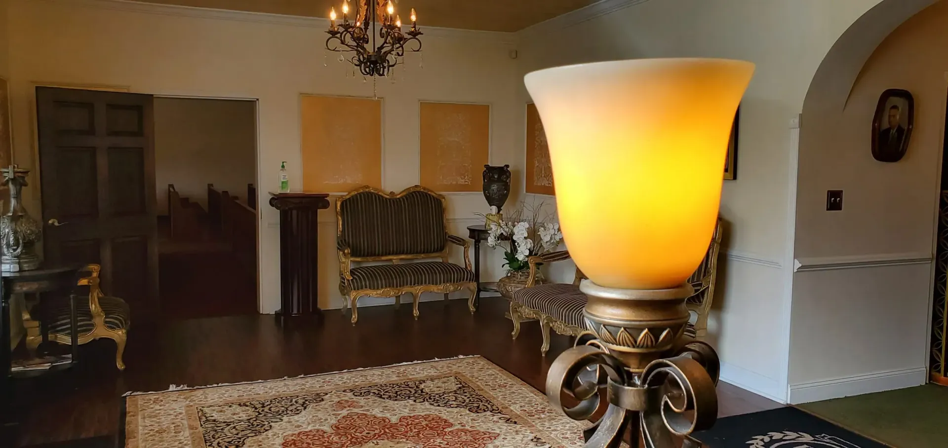 A lit, ornate table lamp with a soft yellow shade in a room with antique furniture.