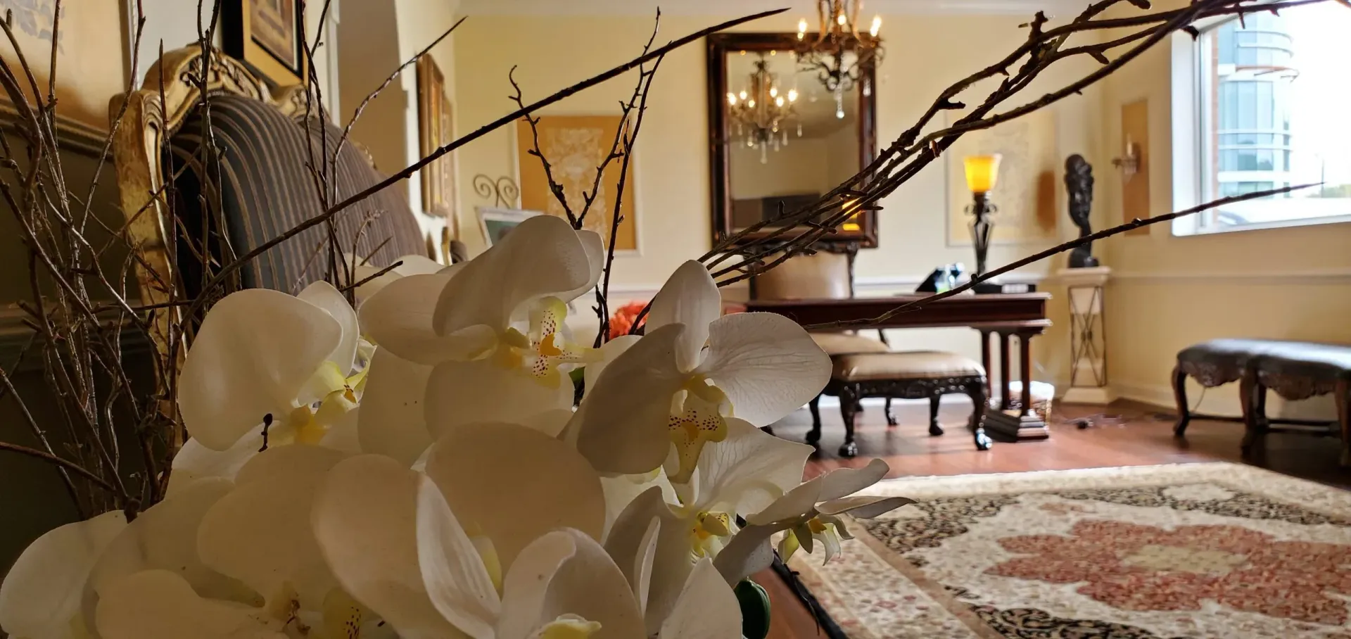 White orchids in a vase, with branches, in a well-lit room with a desk, mirror, and oriental rug.