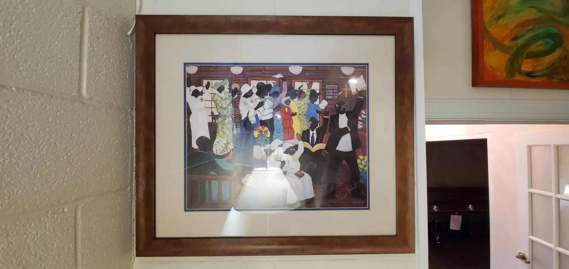 Framed artwork of a colorful gathering; a bride in white dances, brown frame on a white brick wall.
