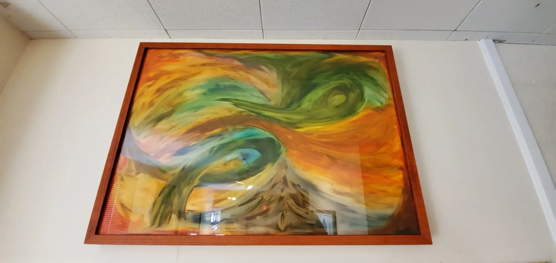 Abstract painting in a wooden frame on a cream-colored wall, featuring swirling green, yellow, and brown colors.