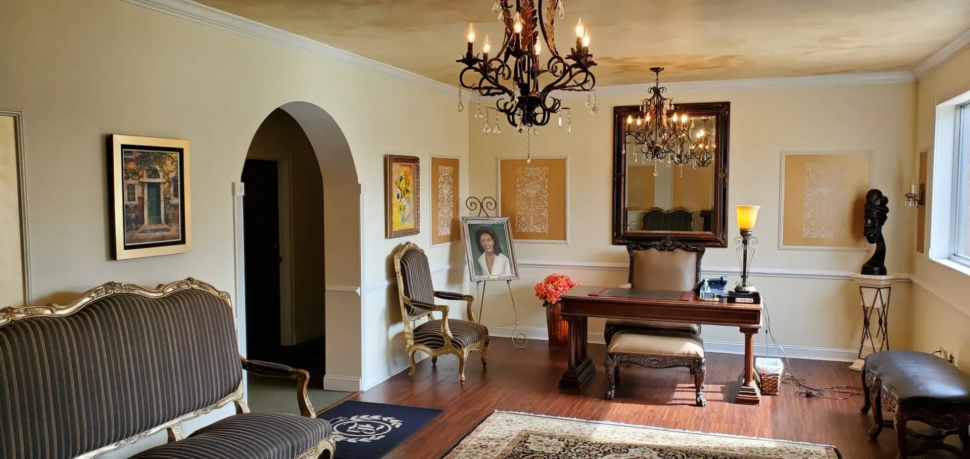 An ornate living room with chandelier, artwork, desk, and antique furniture.