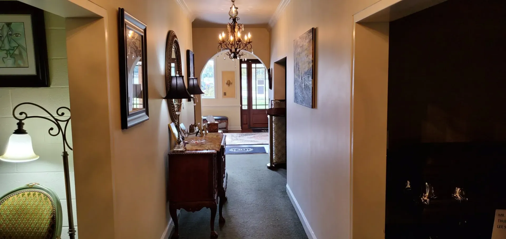 Hallway with antique furniture, art, and chandelier, leading to a doorway.