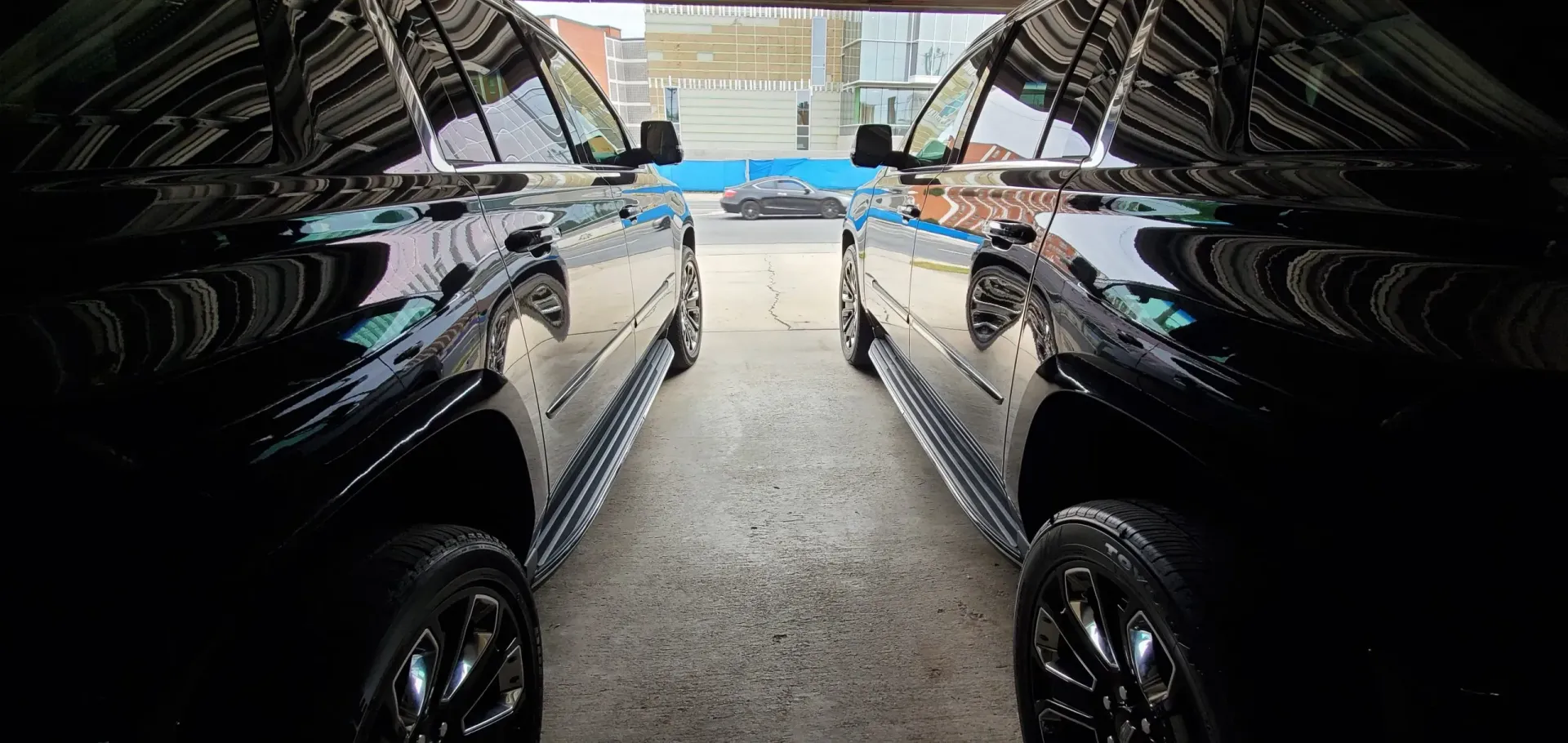Two black trucks parked side-by-side, viewed from between them. They are in a garage or similar area.