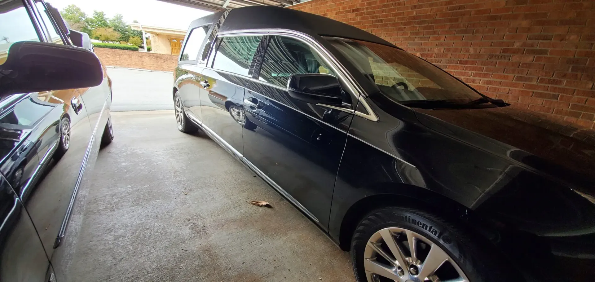 A black hearse parked under a shelter next to a black car, both on a concrete surface.