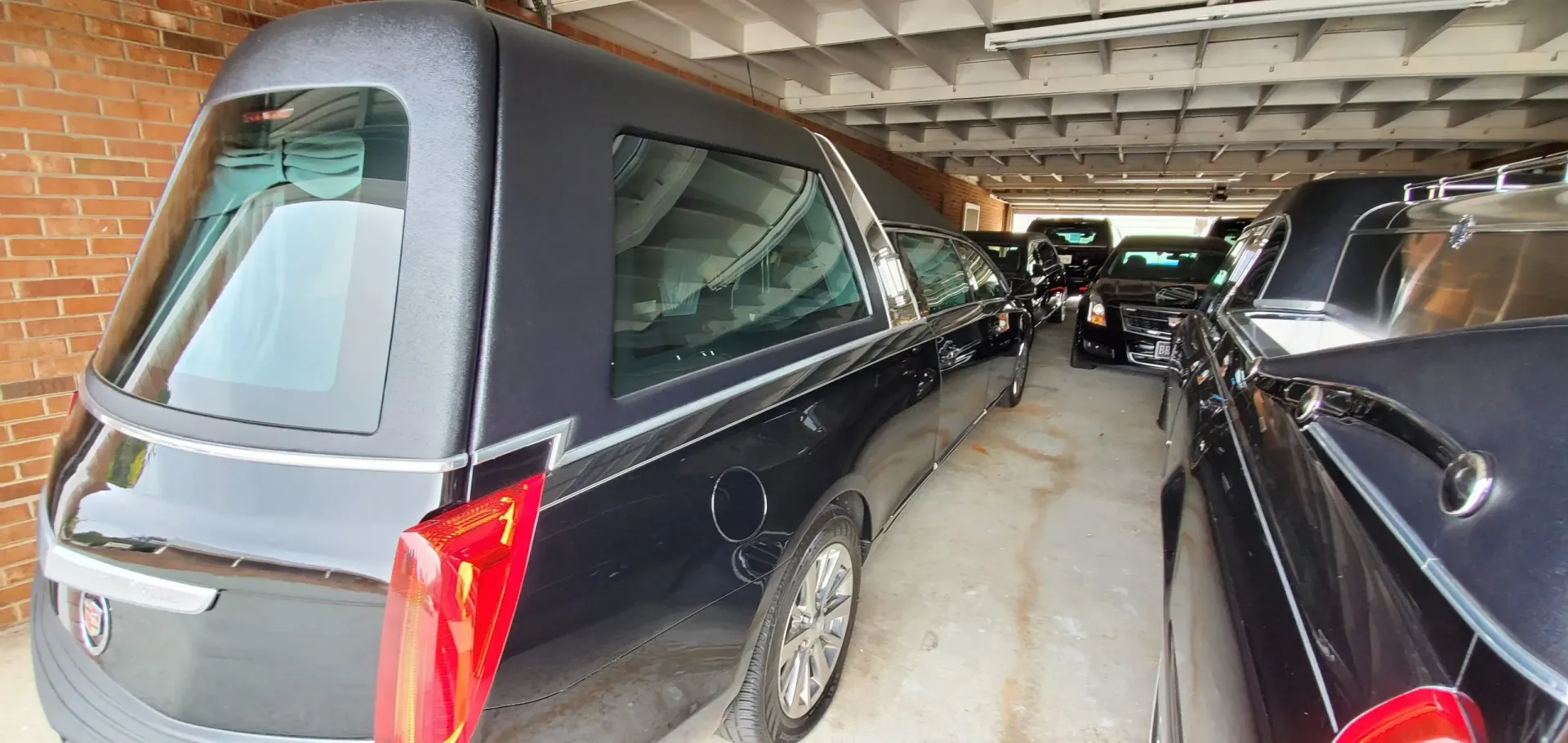 Black hearse parked in a garage with other black vehicles. The garage has a brick wall.