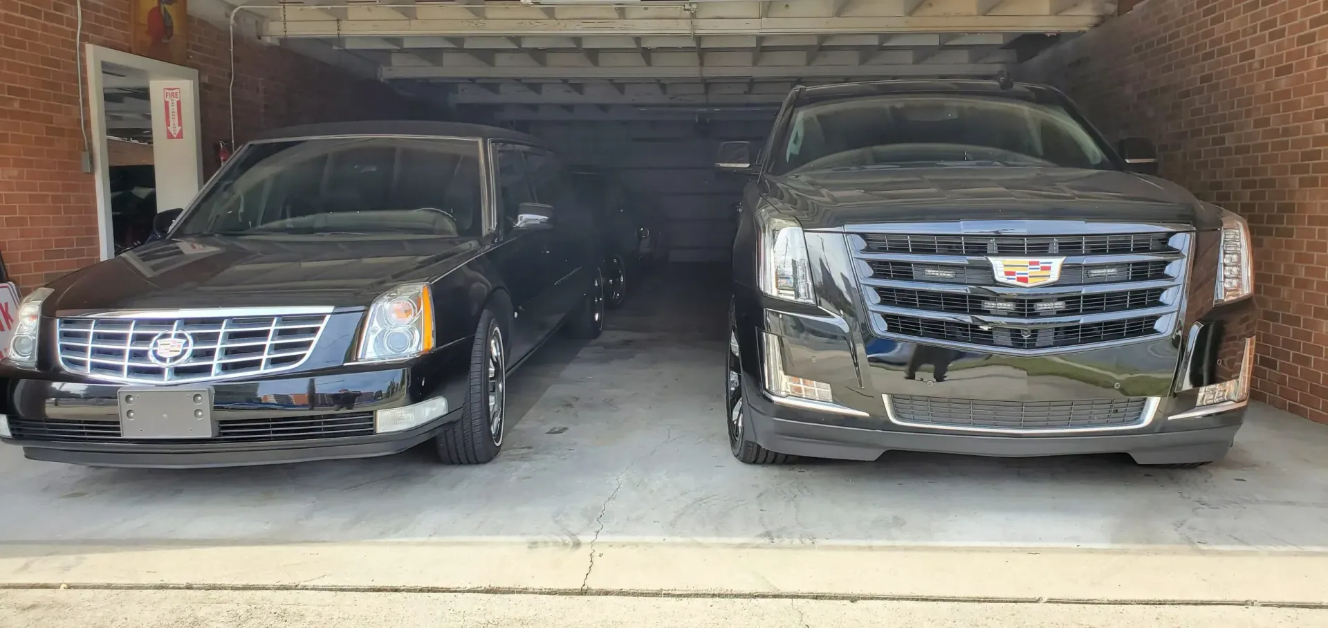 Two black Cadillac vehicles parked inside a garage, with brick walls and a metal ceiling.