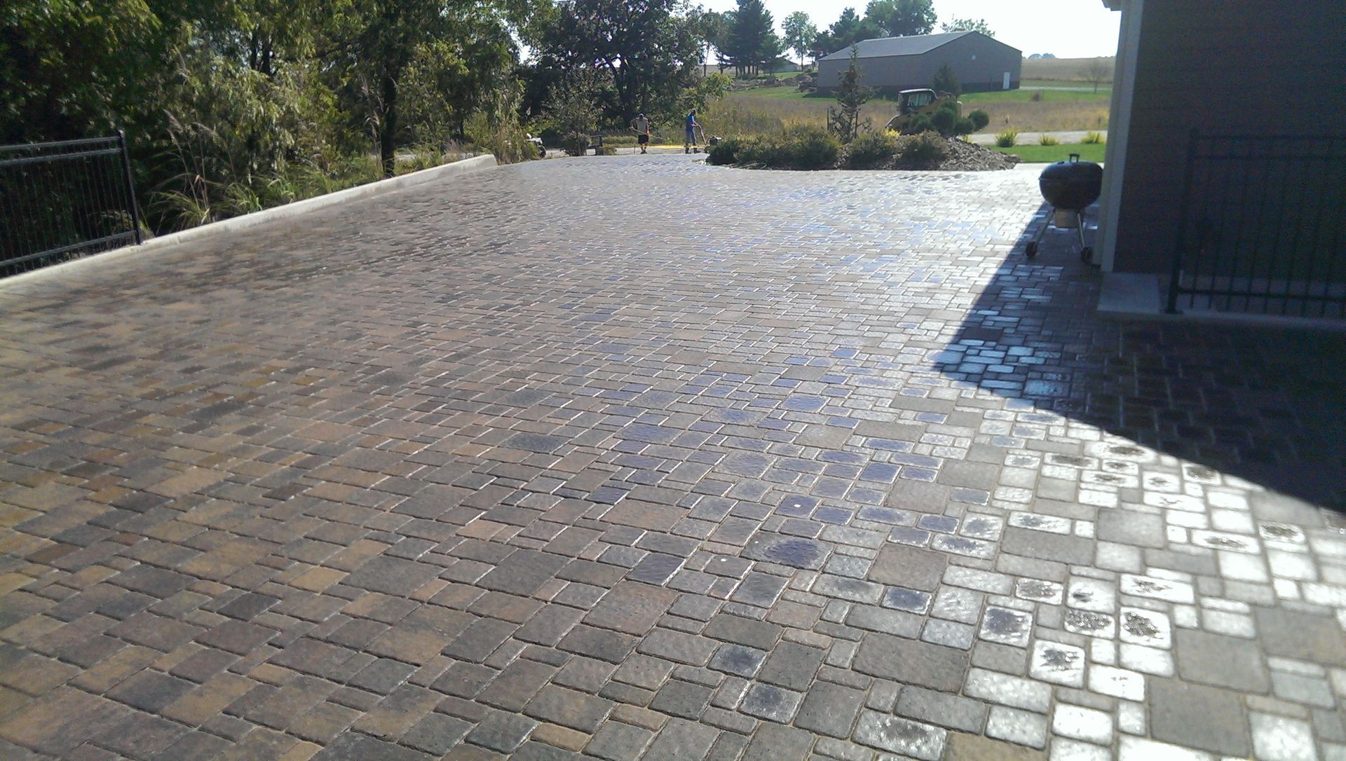 Brick paver driveway in sunlight with a house and trees in the background.