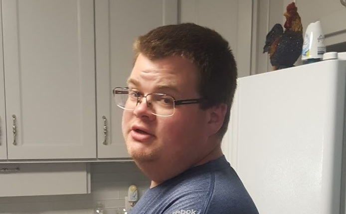 Tick on a green blade of grass, brown body with patterned head, reddish-brown legs, against a blurred green background.