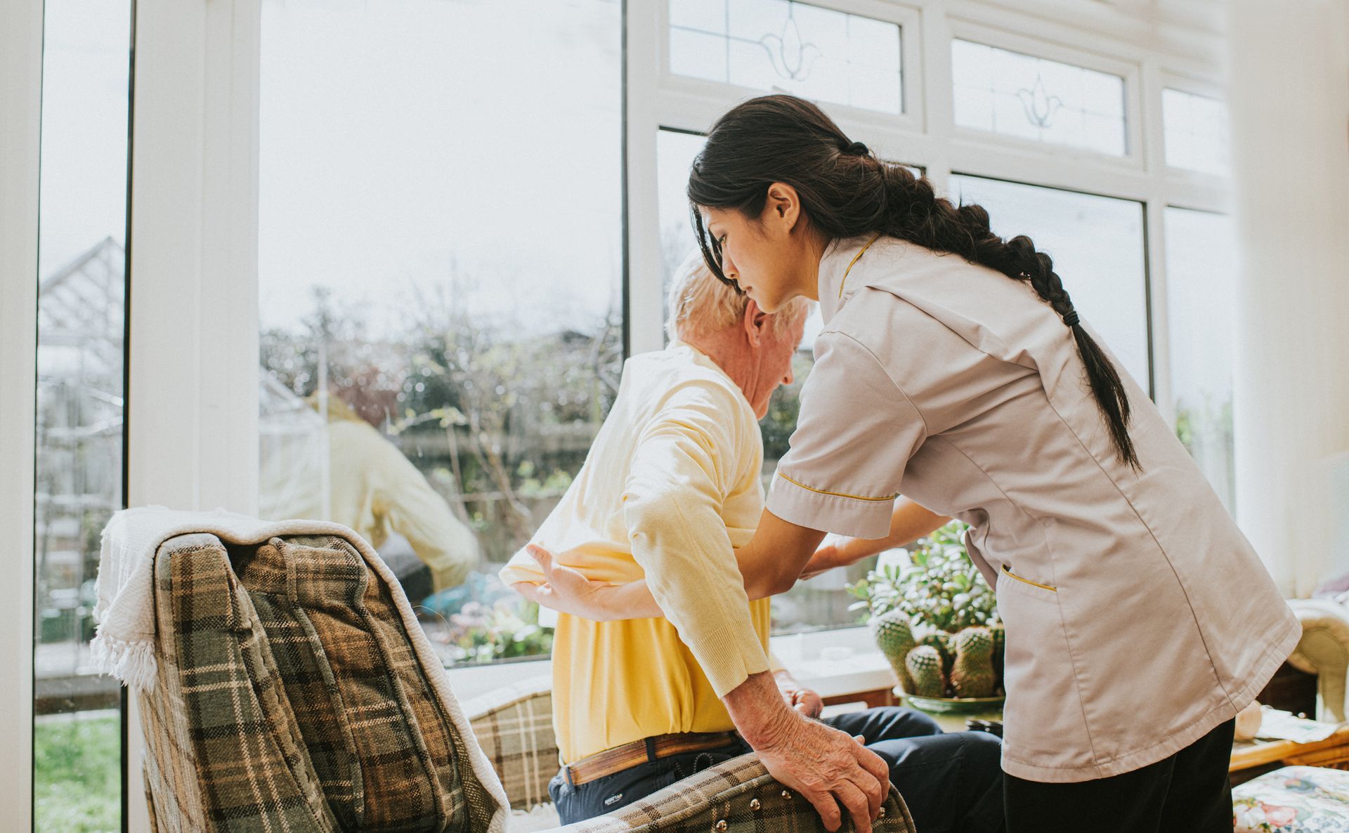 Caregiver assisting a person in sitting up from an armchair, indoors, near a window.