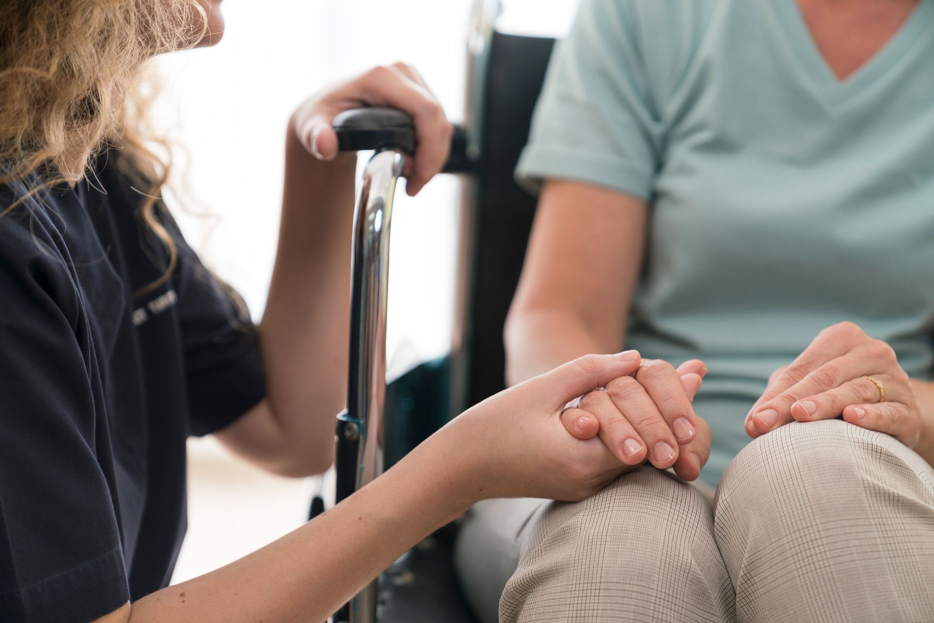 Person in wheelchair holding hands with a caregiver, indoors. Both are wearing neutral-colored clothing, offering comfort.