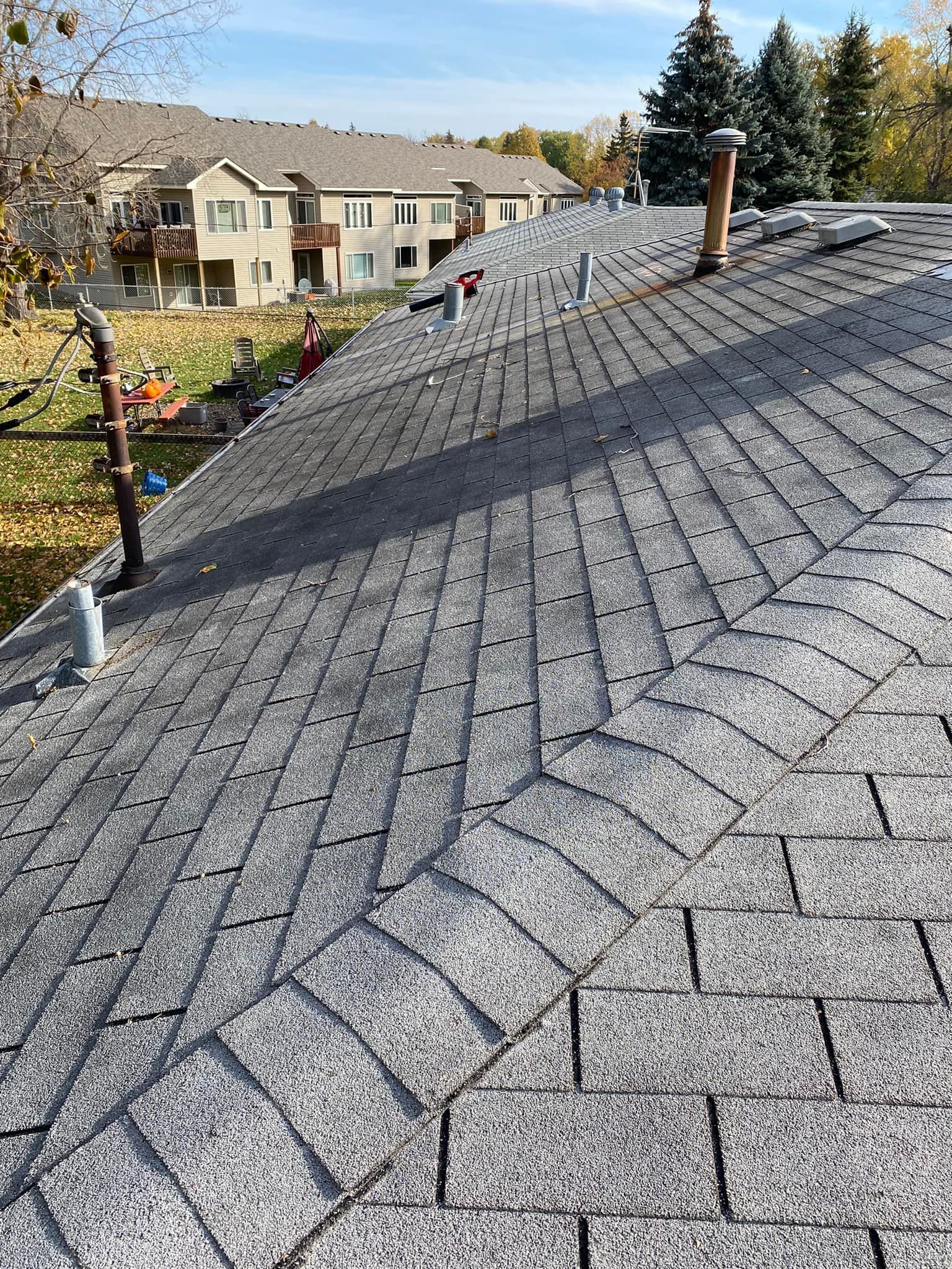 Gray shingled roof with a curved design, with chimneys, pipes, and buildings in the background.