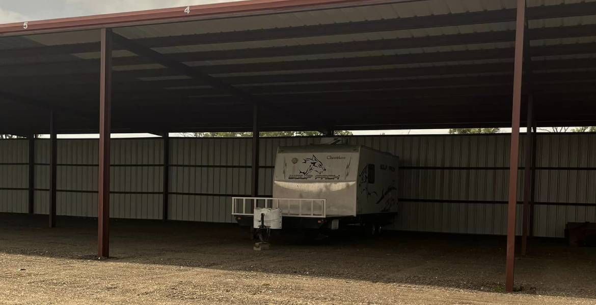 A camper trailer parked under a metal carport, with a gravel ground and corrugated metal fencing.