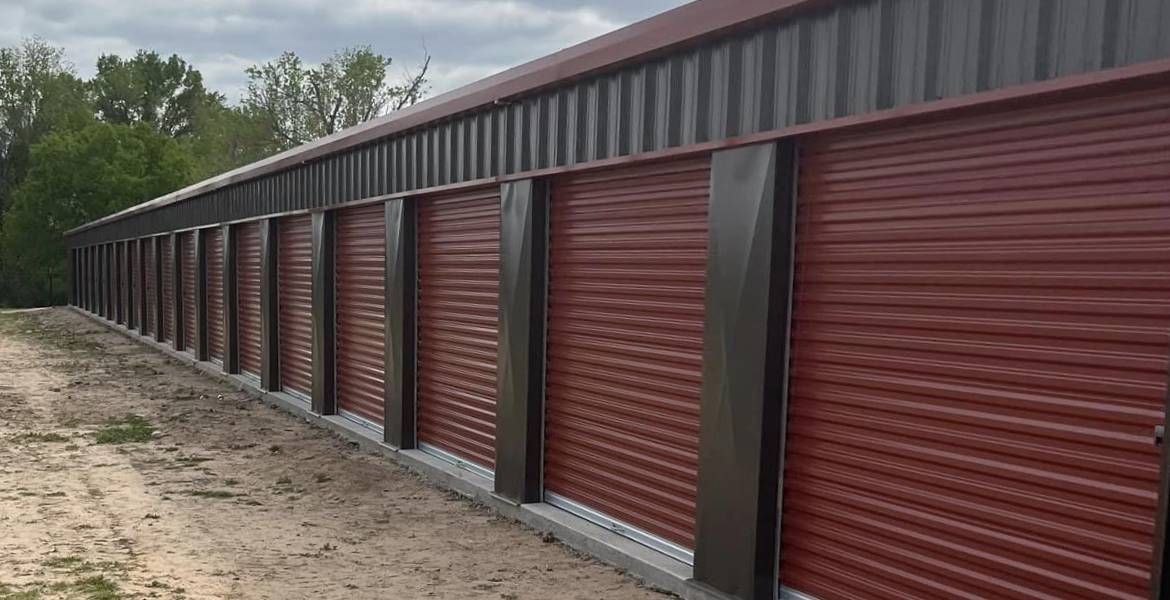 Row of red storage unit doors in a gray metal building, set in a dirt lot near trees.