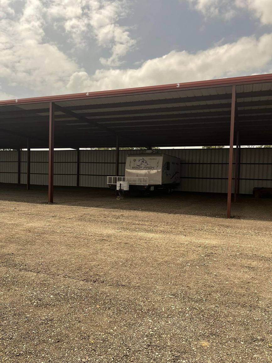 A camper trailer parked under a large metal awning in a gravel lot on a cloudy day.