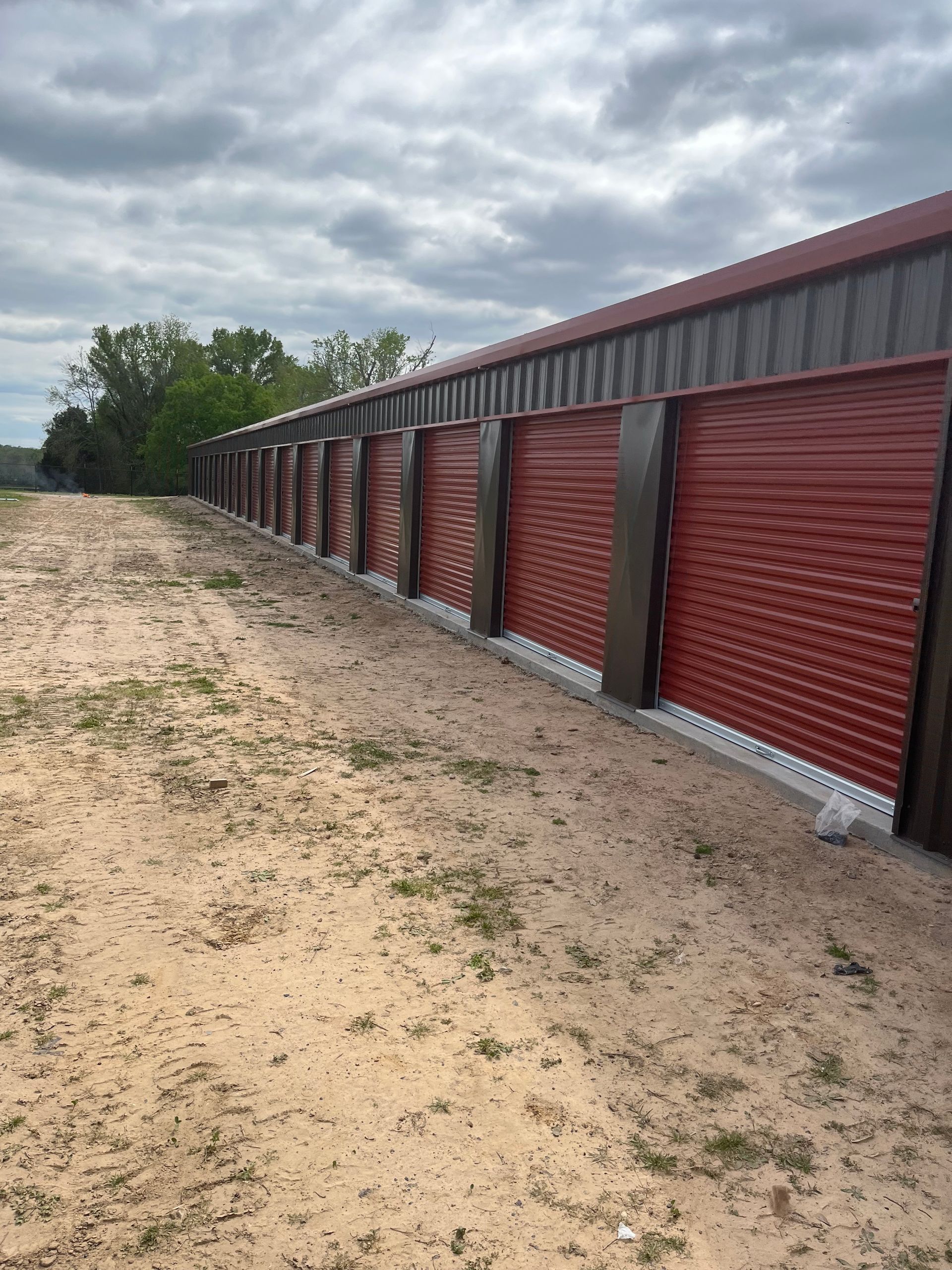 Row of red storage unit doors on a sandy lot, under a cloudy sky.