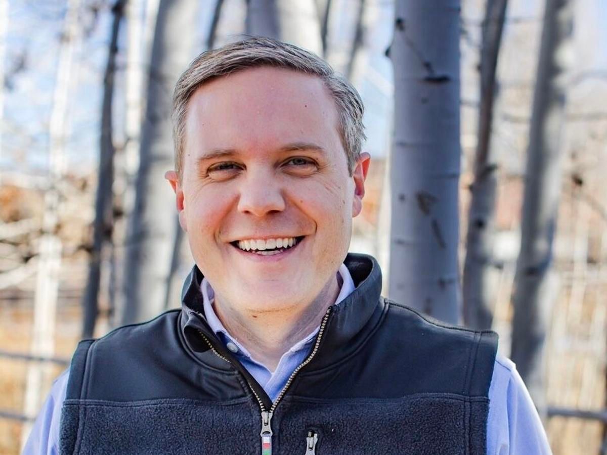 A man wearing a black vest is smiling in front of trees.