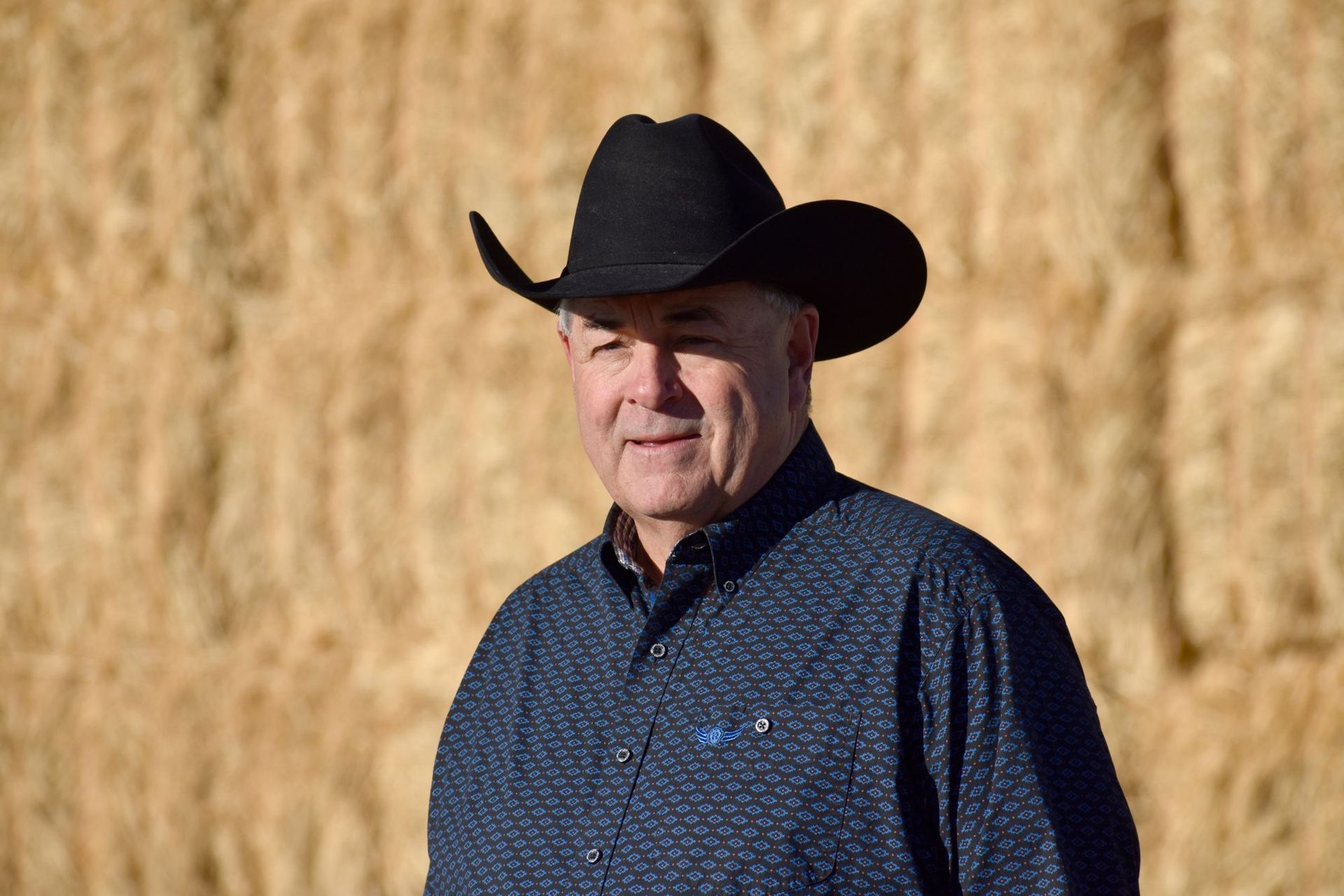 A man wearing a cowboy hat and a blue shirt is standing in front of a pile of hay.