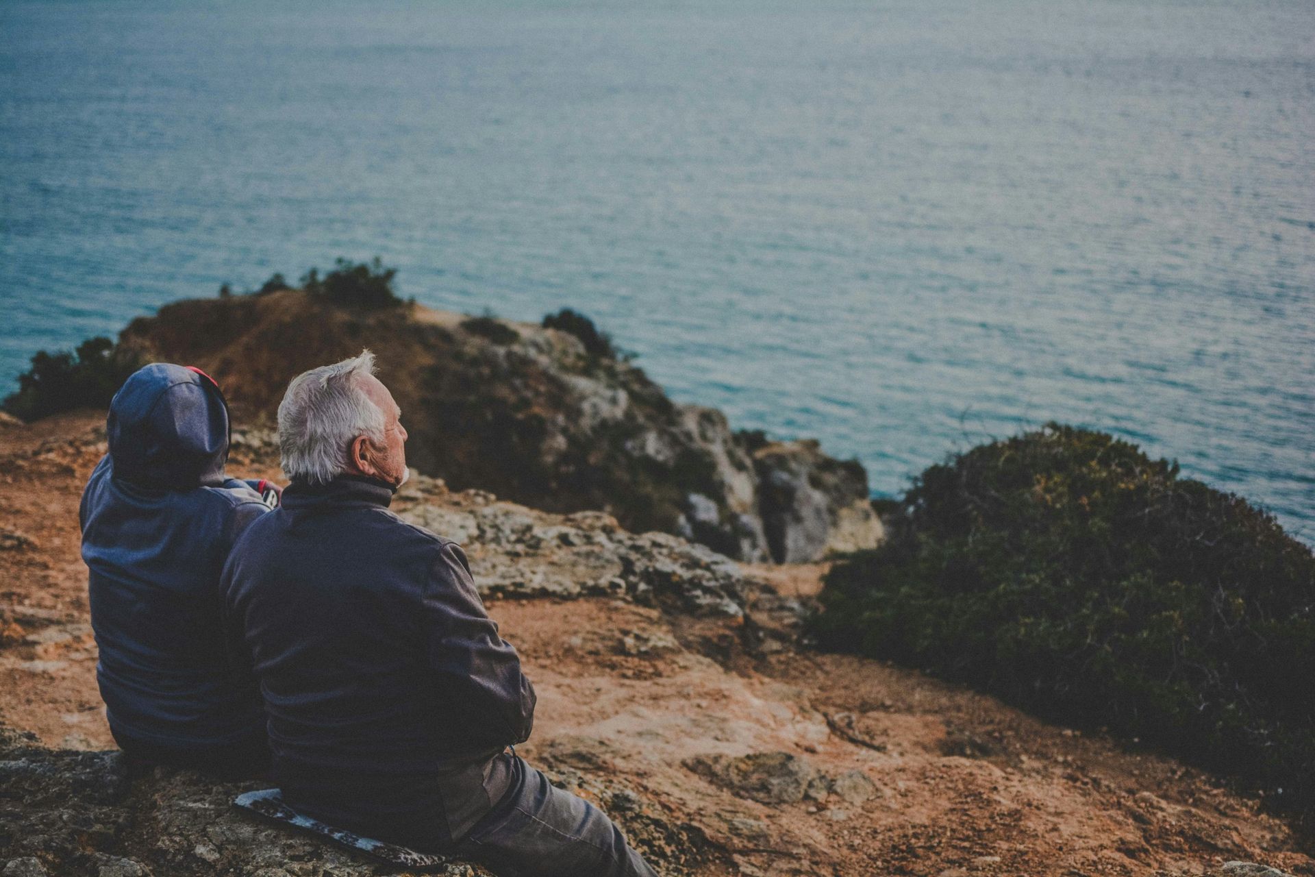 A man and a woman are sitting on top of a cliff overlooking the ocean.