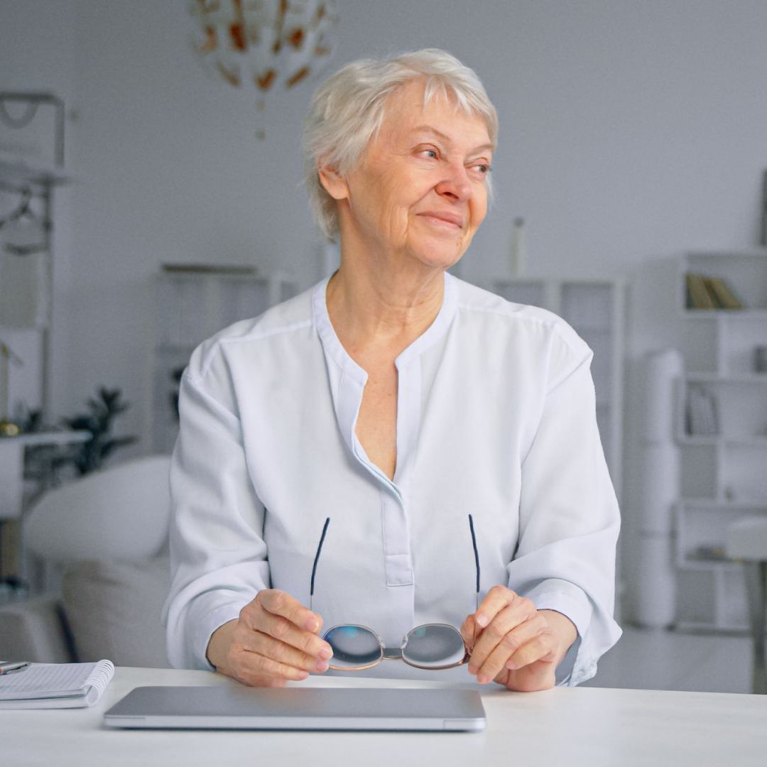 An elderly woman is sitting at a table holding a pair of glasses.