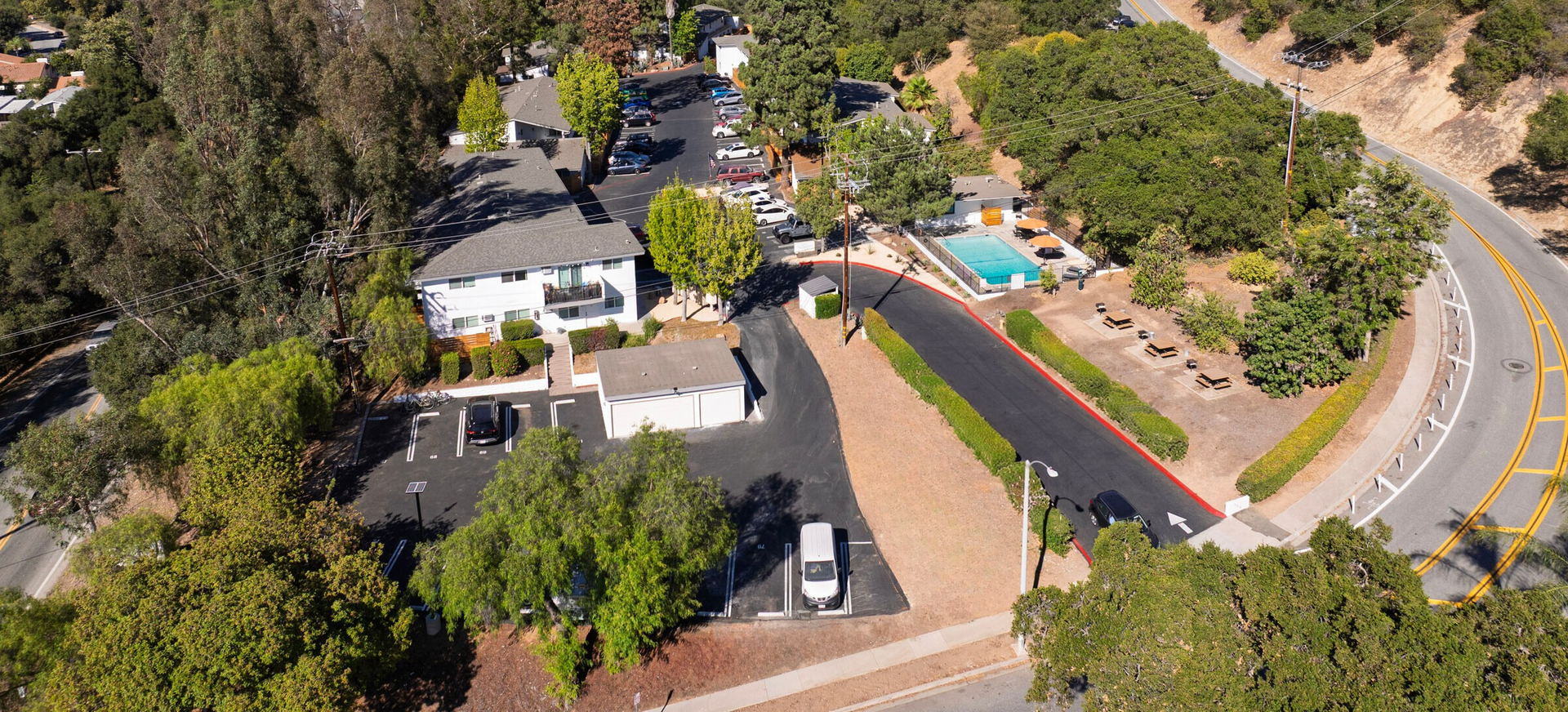 Aerial view of a residential complex with a swimming pool, trees, and a winding road against a backdrop of mountains.