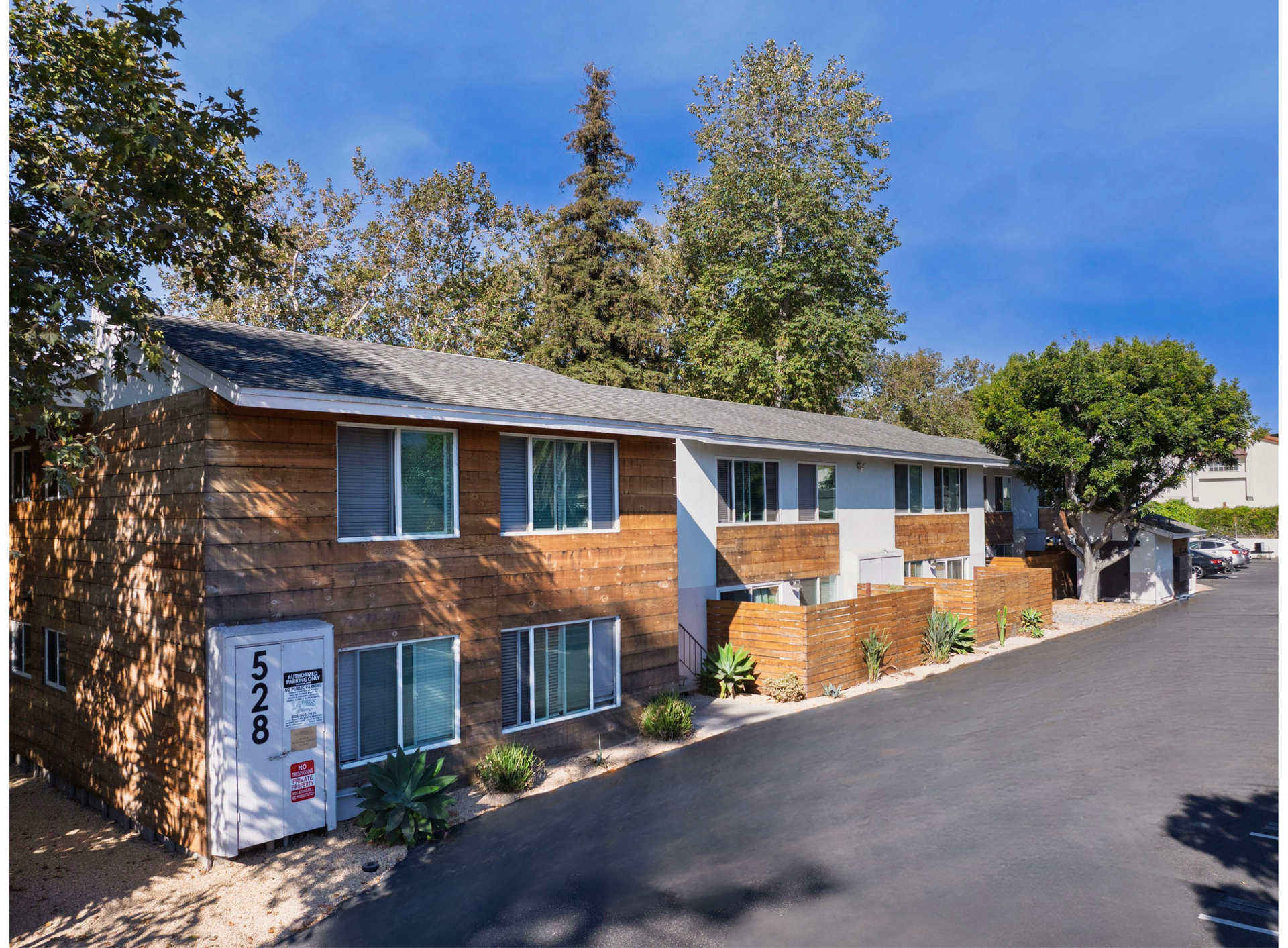 Two-story apartment building with brick accents and a grey roof, set against a background of trees.