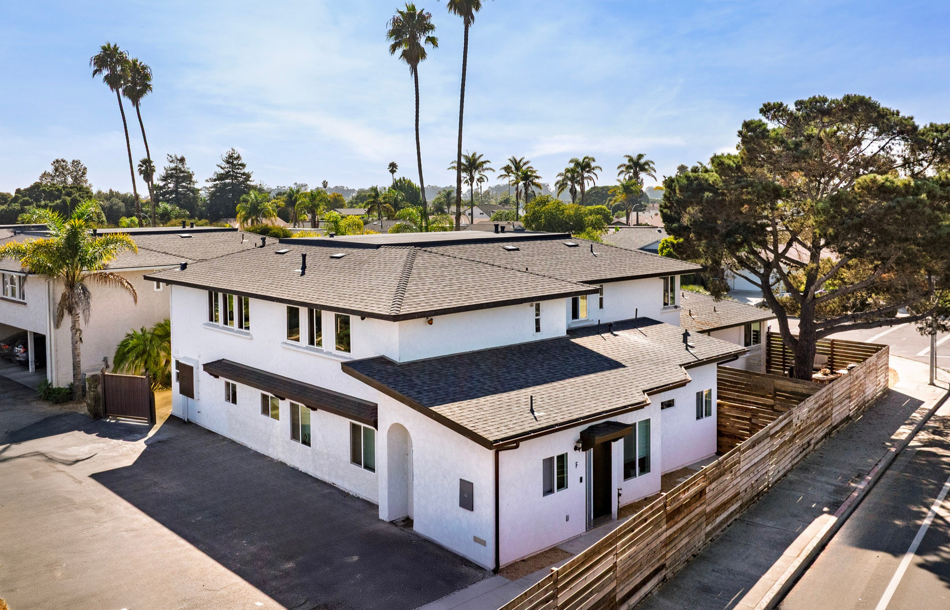 White two-story building with dark roof tiles, palm trees in the background, wooden fence in the foreground.