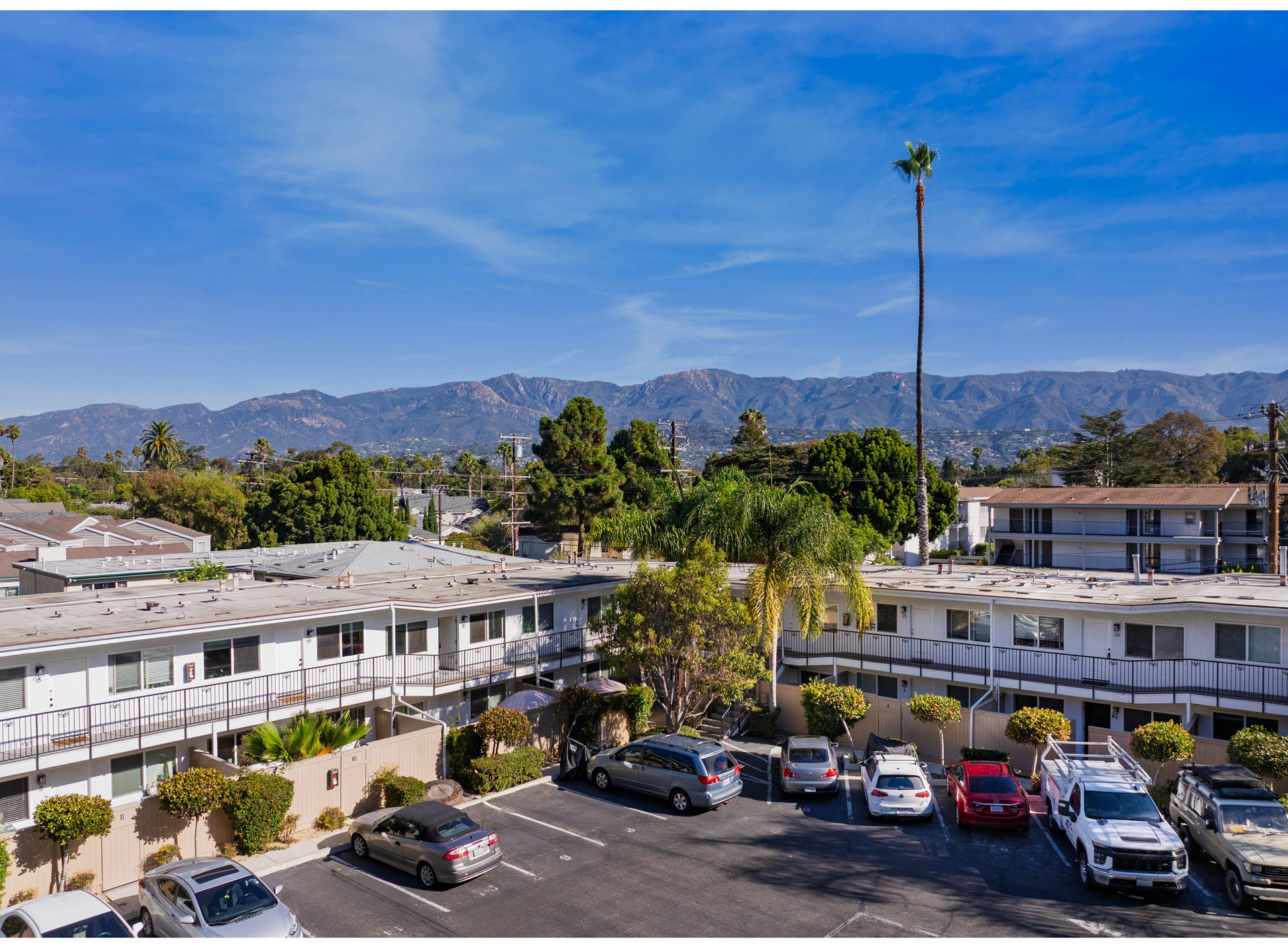 Apartment complex with cars parked in front, trees, and mountains in the background under a blue sky.