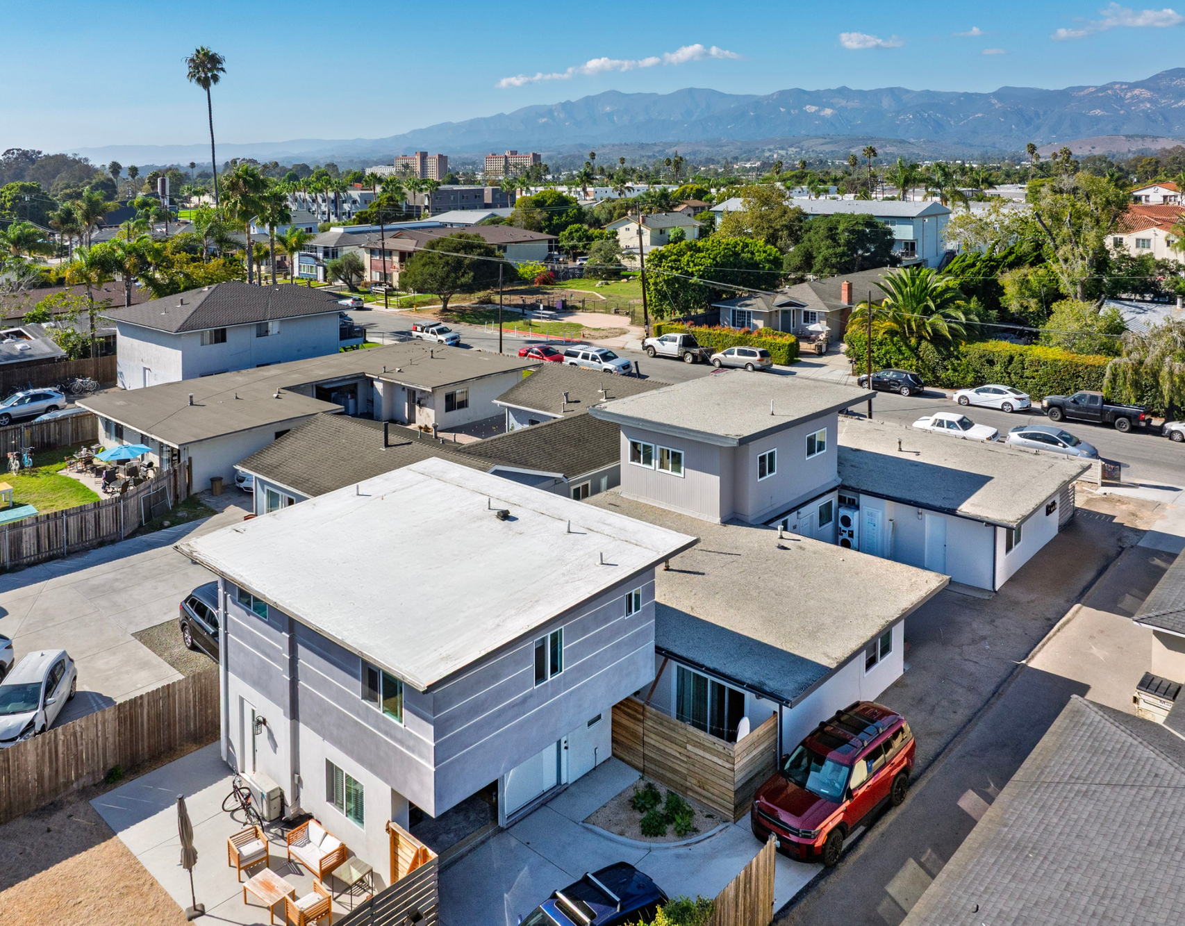 Aerial view of houses with grey roofs and walls, parked cars, trees, and mountains in the background under a blue sky.