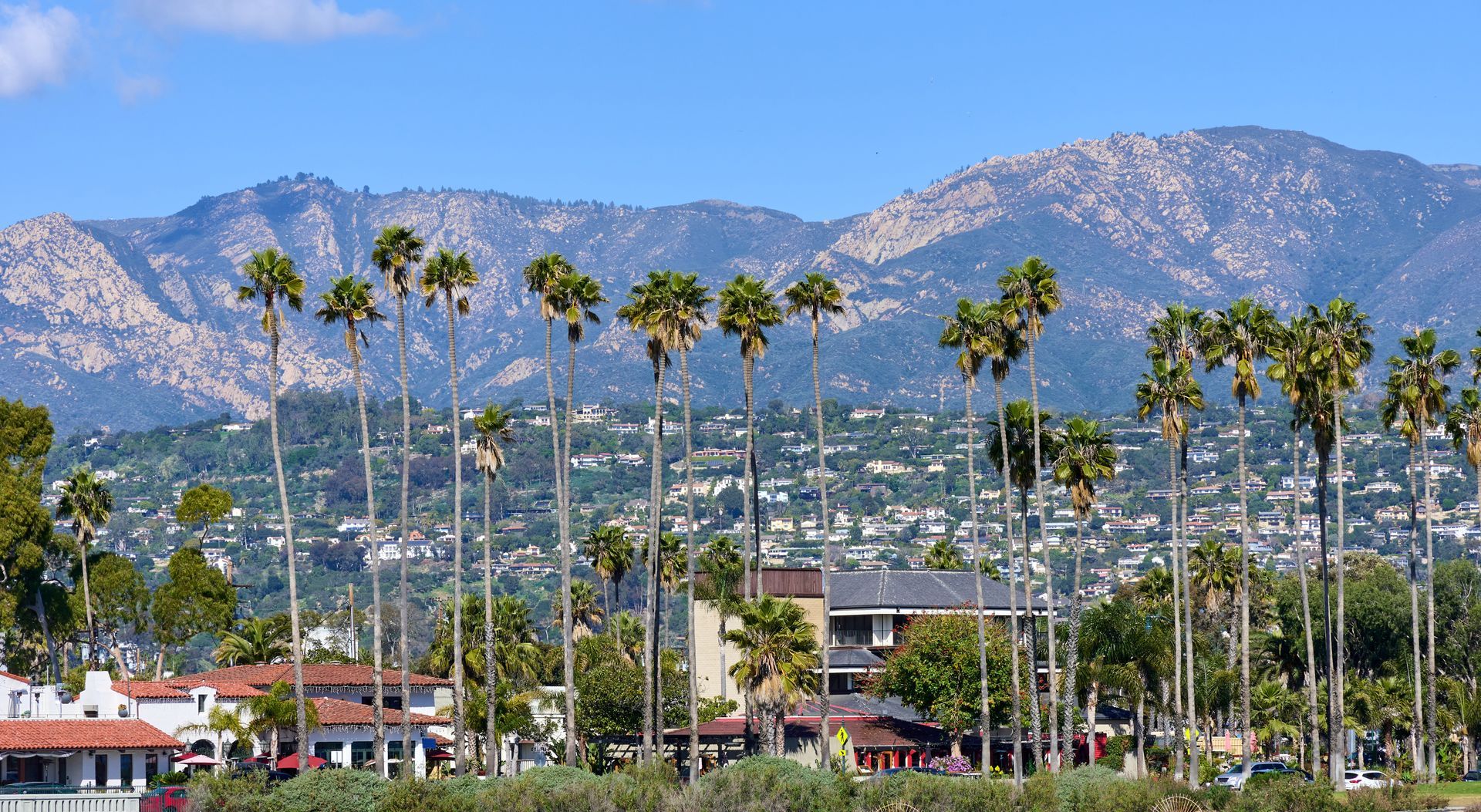 Beachfront scene with buildings, shoreline, ocean waves, and mountains under cloudy skies.