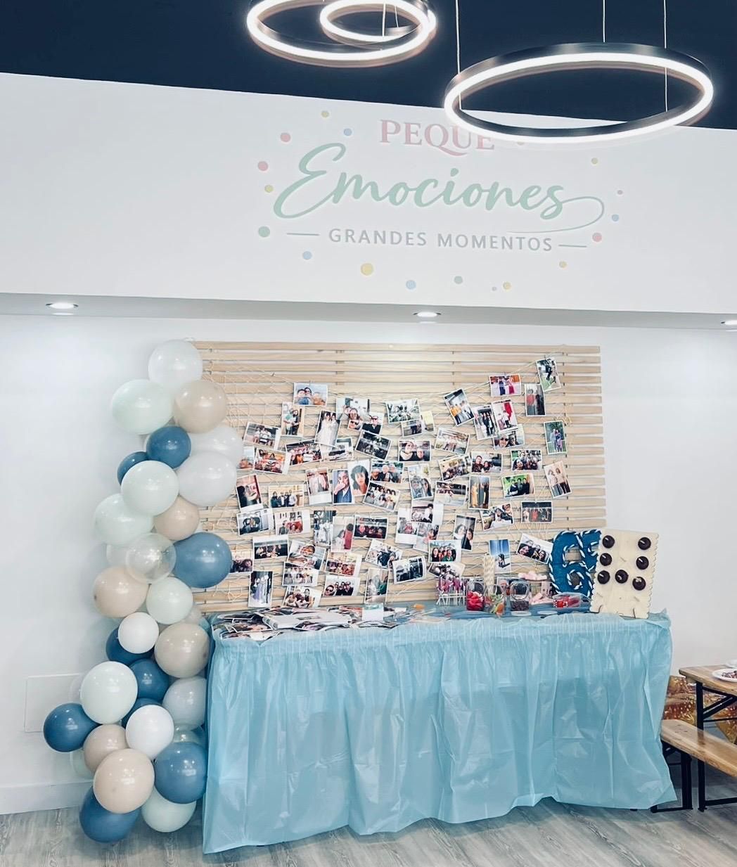 Fondo decorado para el evento con globos, pared con collage de fotos y mesa cubierta con un mantel azul bajo luces de anillo.