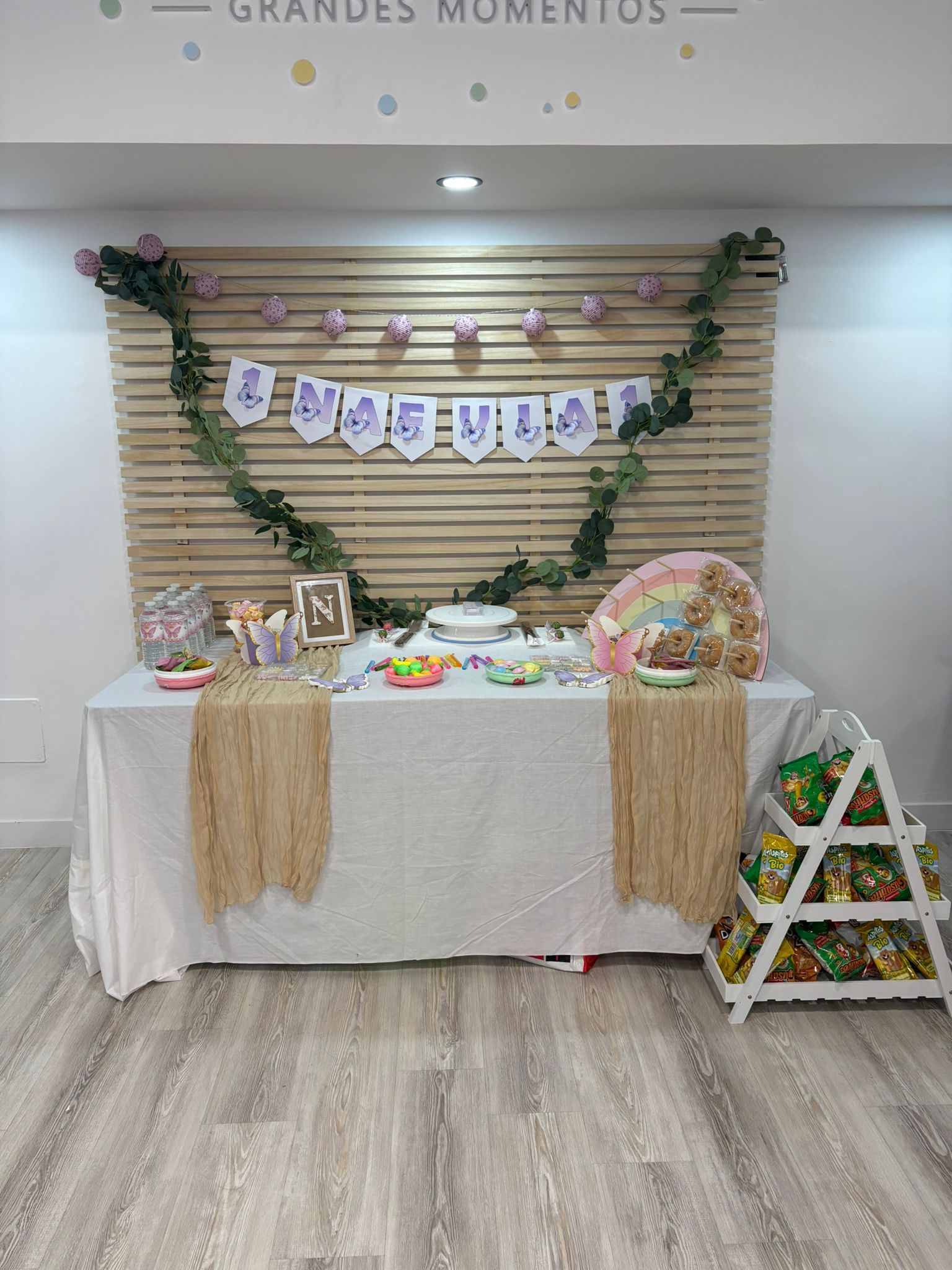 Mesa de postres decorada para el evento con mantel blanco, guirnalda floral, pancarta y una pequeña escalera a la derecha.