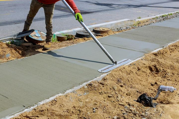 Worker smoothing a fresh concrete sidewalk surface near a road.