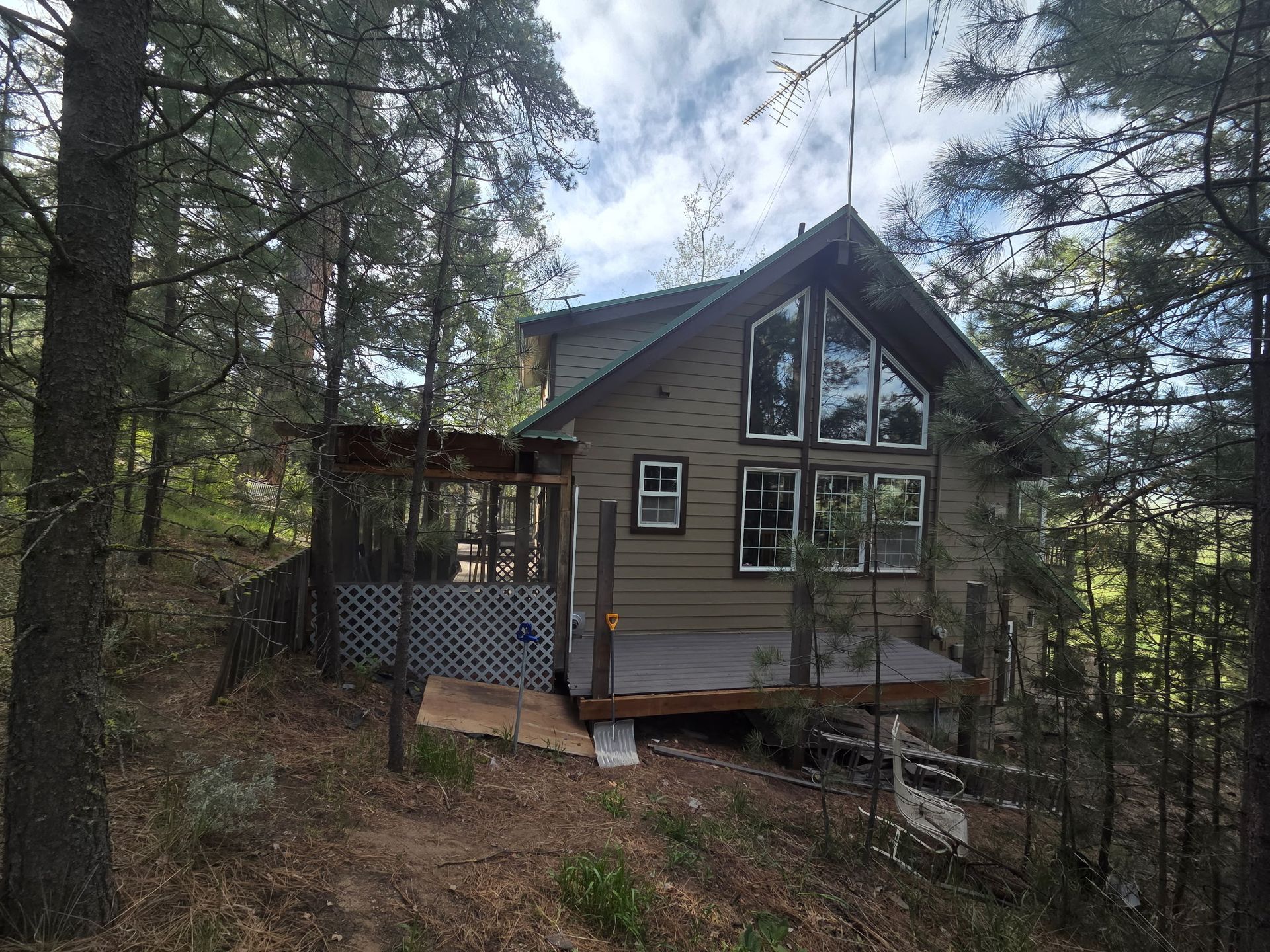 Small gray cabin with large windows on a wooded hillside, surrounded by tall pine trees.