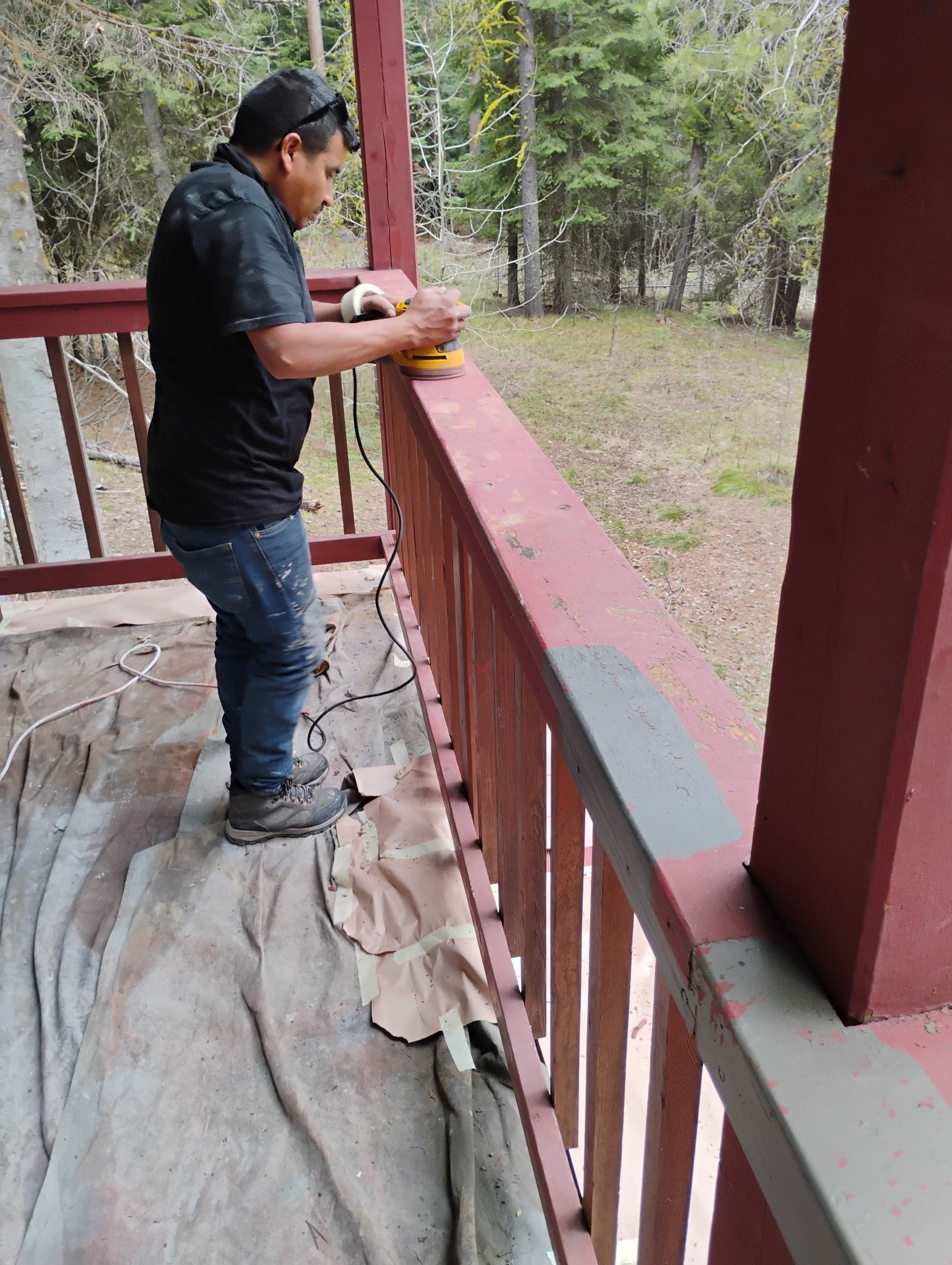 Person standing on a porch railing, painting it red outdoors near a wooded area.